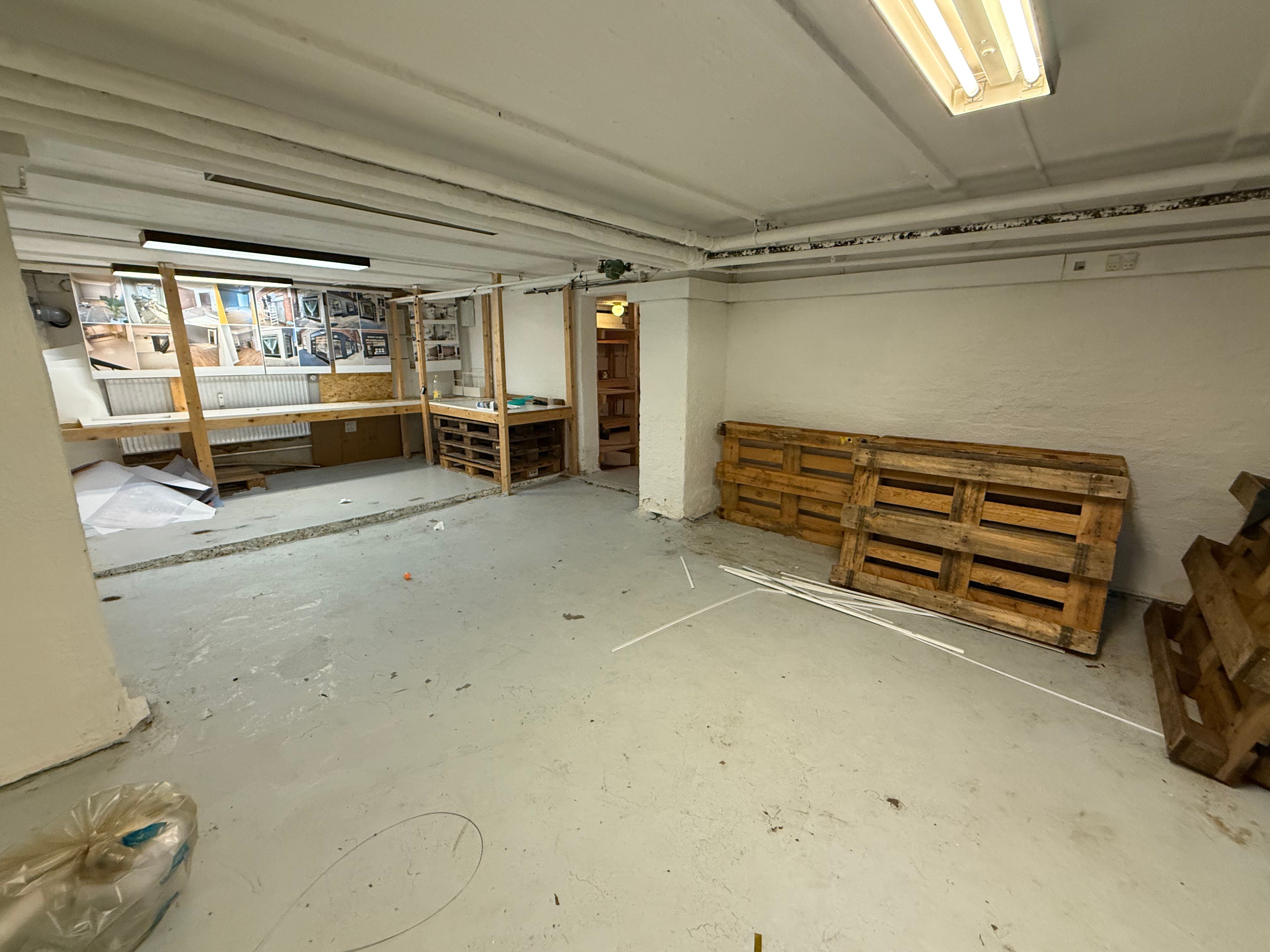 Lagerrum med to høje af trækasser, to arbejdsborde og to lysekroner samt tøj i en plastikpose på gulvet