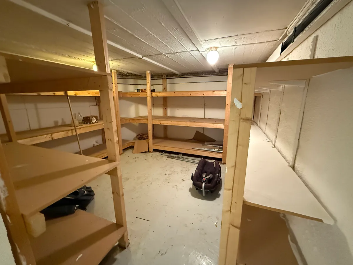 Værelse med lysegule vægge og hvide lofter med to rækker træhylder og et barnesæde på gulvet.