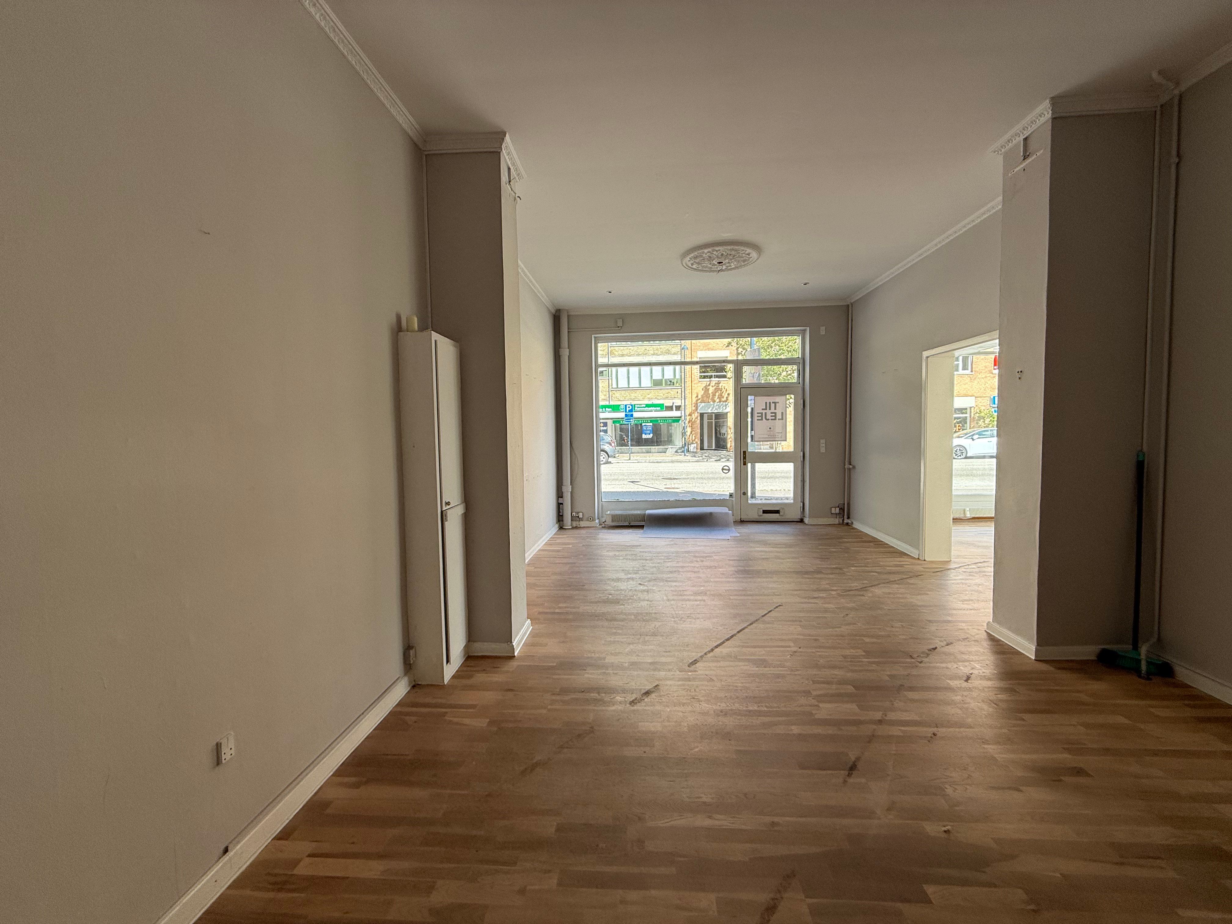 En tom og ren butik med lyse trægulve og hvide vægge og døre, der vender mod en åben indgang