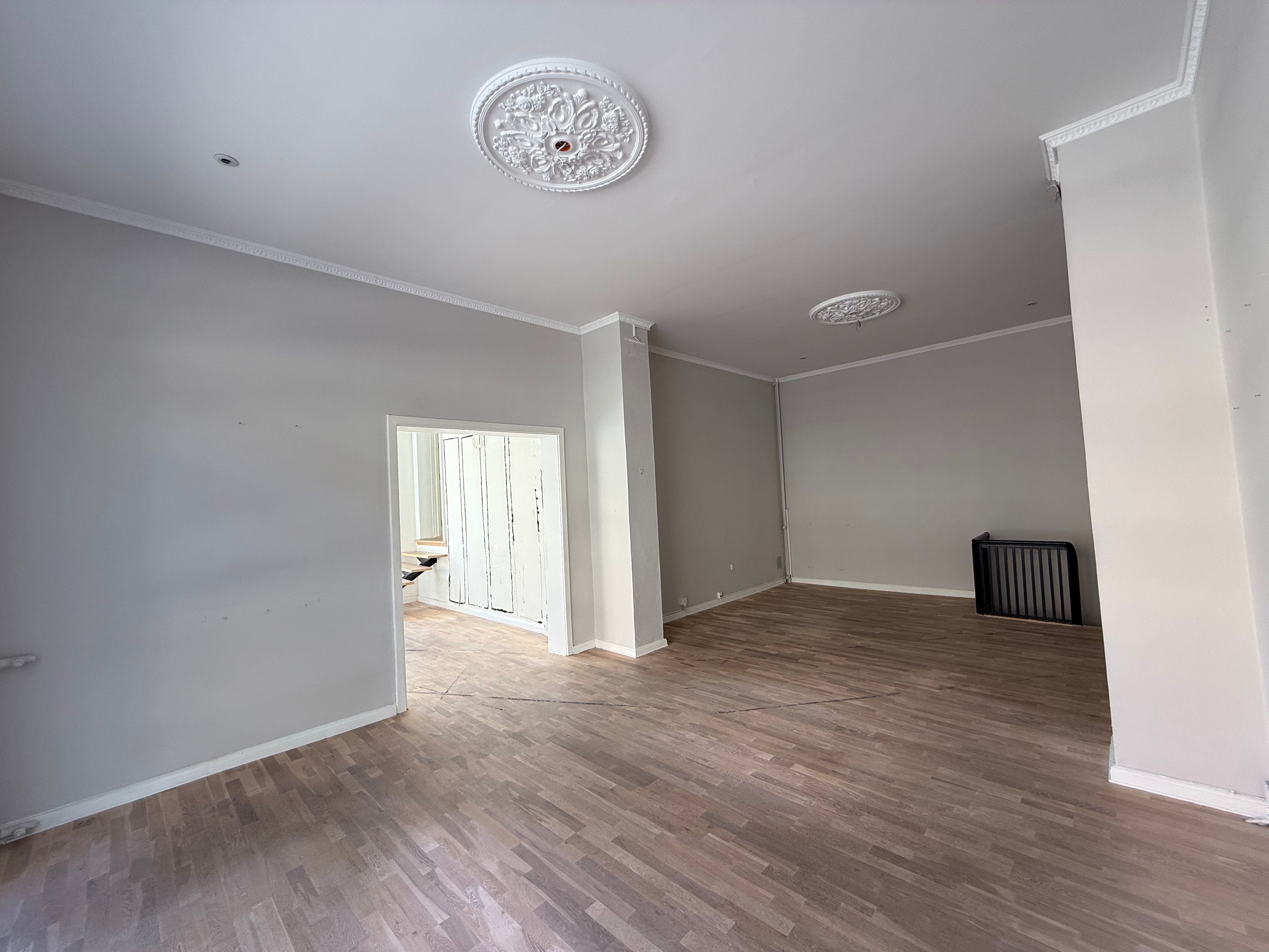 Et tomt rum med høje lofter og hårdt trægulv, to store lyseplader og en hvid søjle på højre side