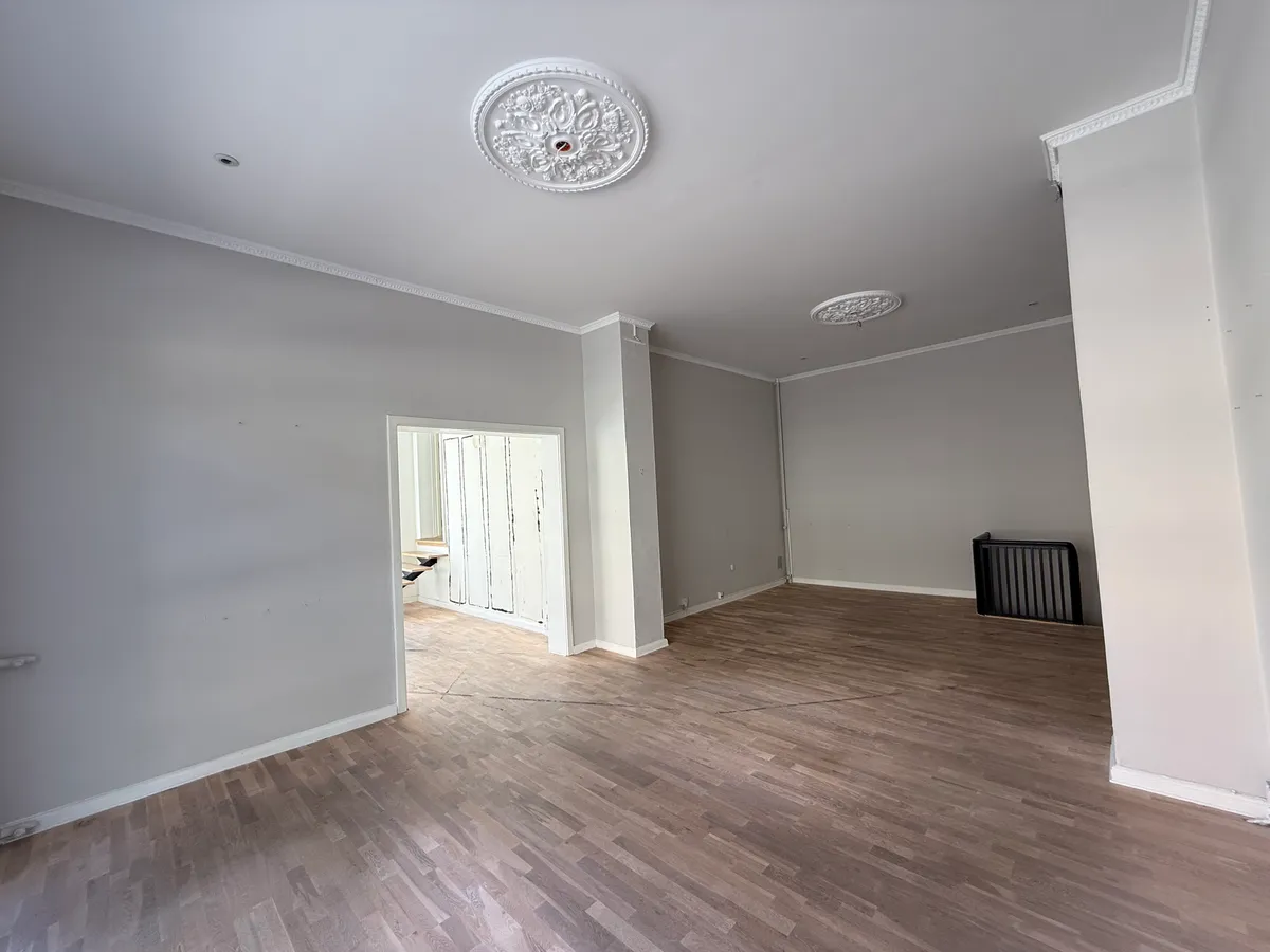 Et tomt rum med høje lofter og hårdt trægulv, to store lyseplader og en hvid søjle på højre side