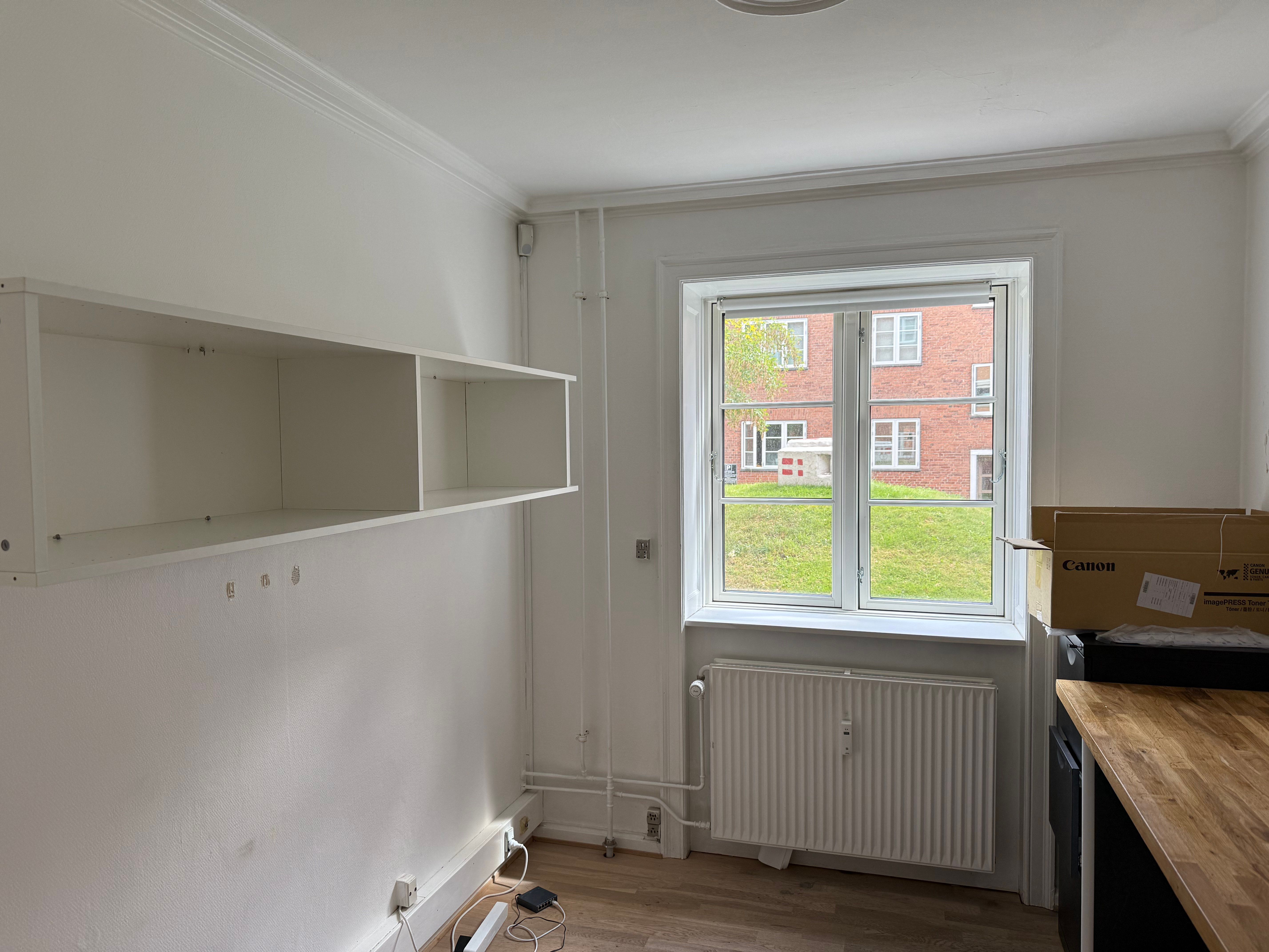 Et tomt rum med hvide vægge, et hjørnehylde og en radiator under vinduet, der ser ud på en grøn park