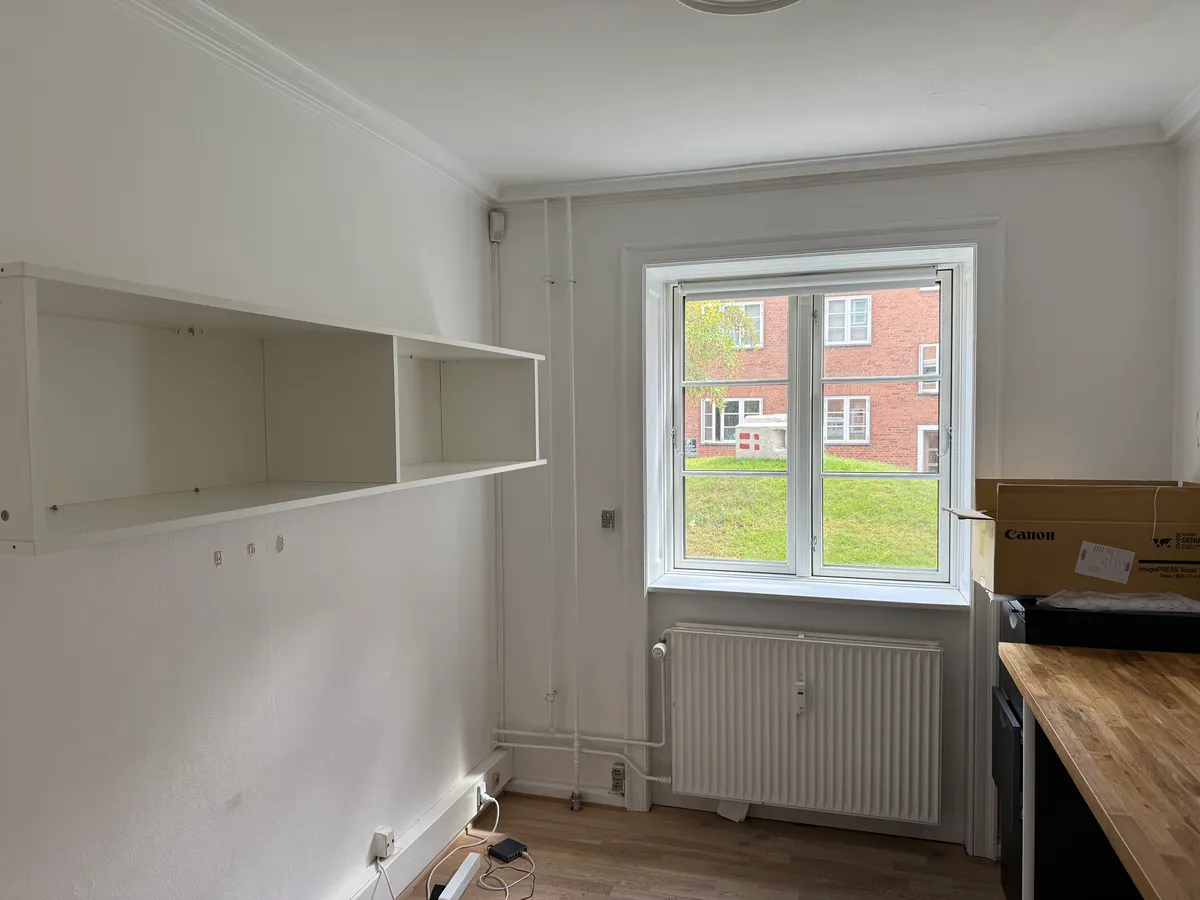 Et tomt rum med hvide vægge, et hjørnehylde og en radiator under vinduet, der ser ud på en grøn park