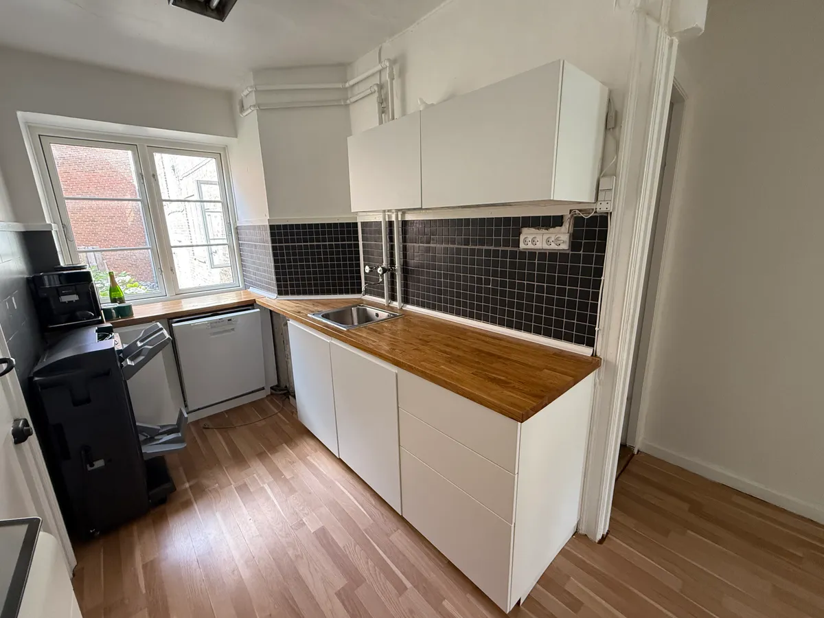 En moderne køkken med hvide vægge, trægulv og højtløftede hvide skabe samt et træbord med en integreret ovn og en sink