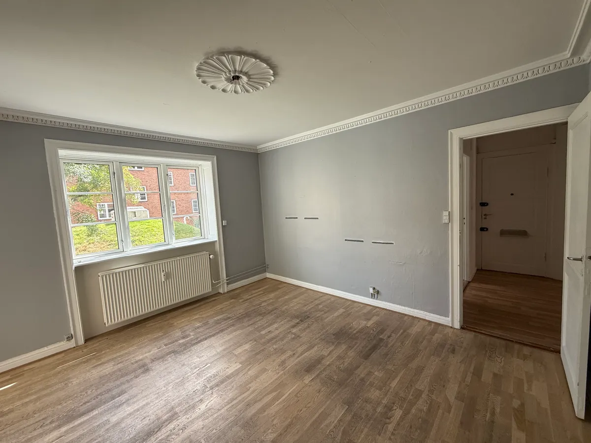 En stor stue med lysegrå vægge og hårdt trægulv og en hvid radiator under vinduet med udsigt til grønne områder