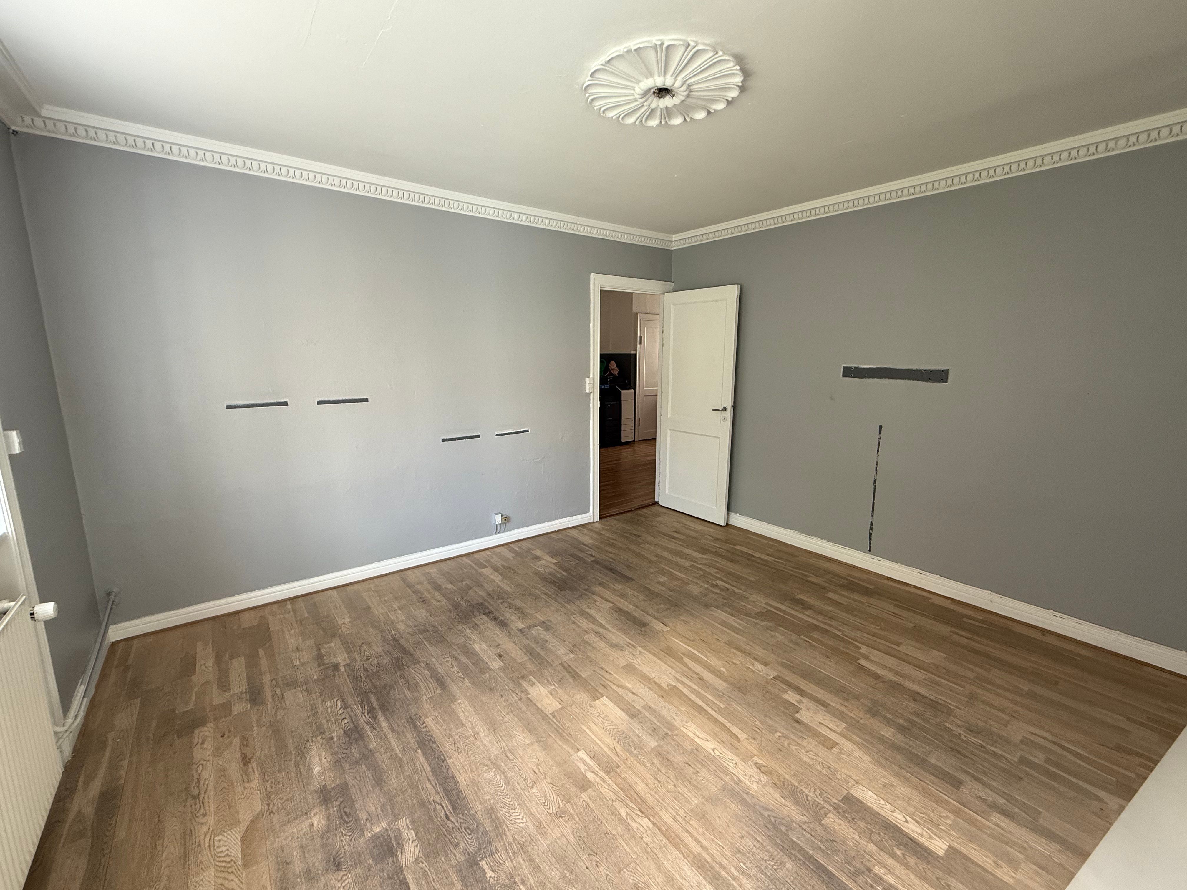 En tom lejlighed med lysegrå vægge og en åben dør til et badeværelse med hvid stryget dør og et vindue til et køkken