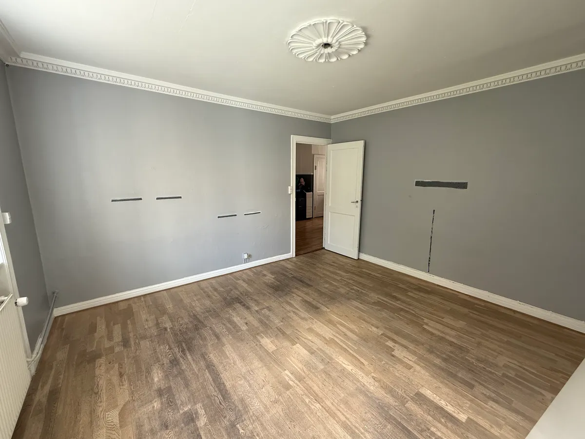 En tom lejlighed med lysegrå vægge og en åben dør til et badeværelse med hvid stryget dør og et vindue til et køkken