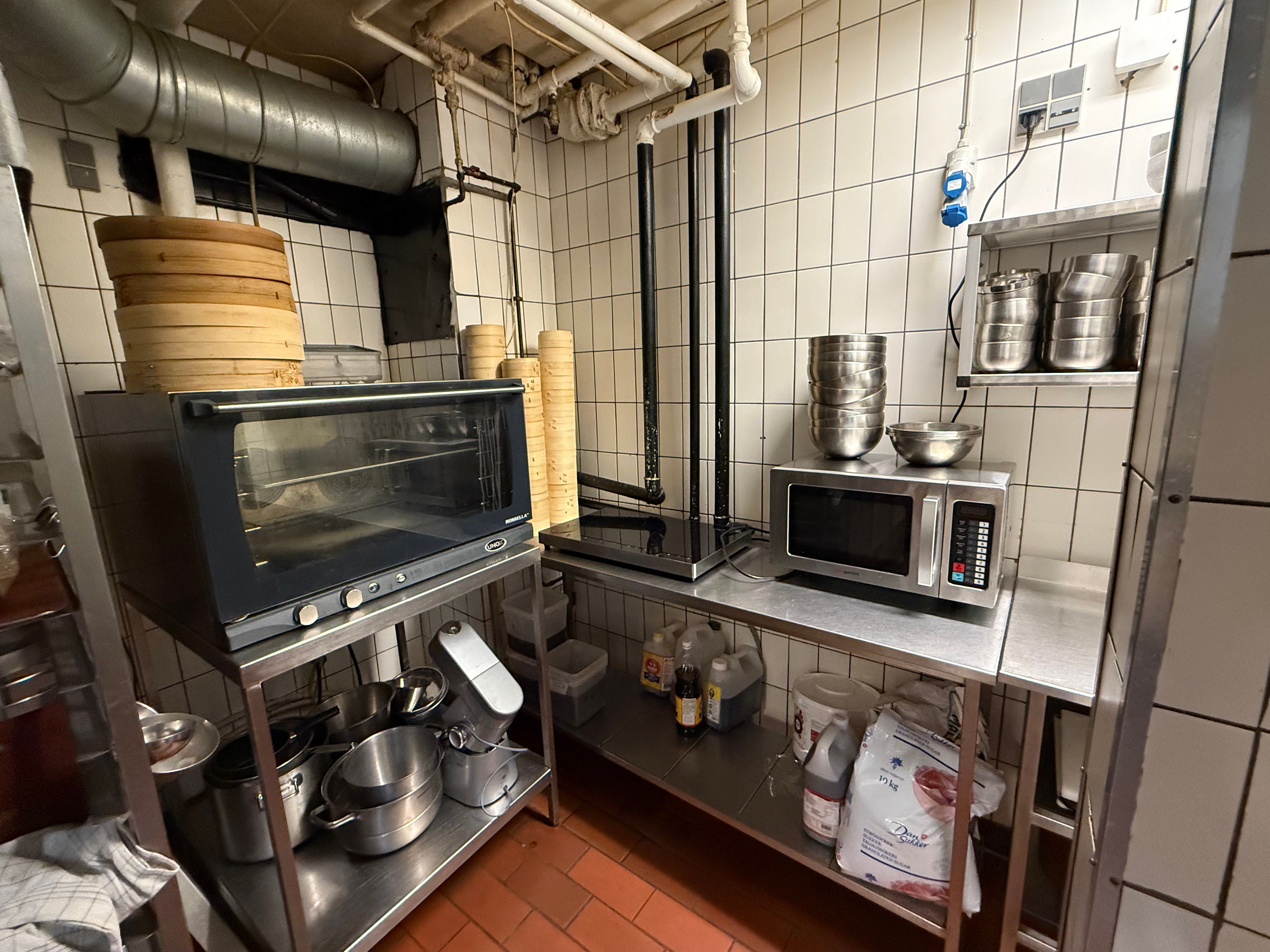 Køkken med komfuret, ovnen, mikrobølgeovnen, flere beholdere, bønner og flasker samt en række metalborde med køkkenudstyr
