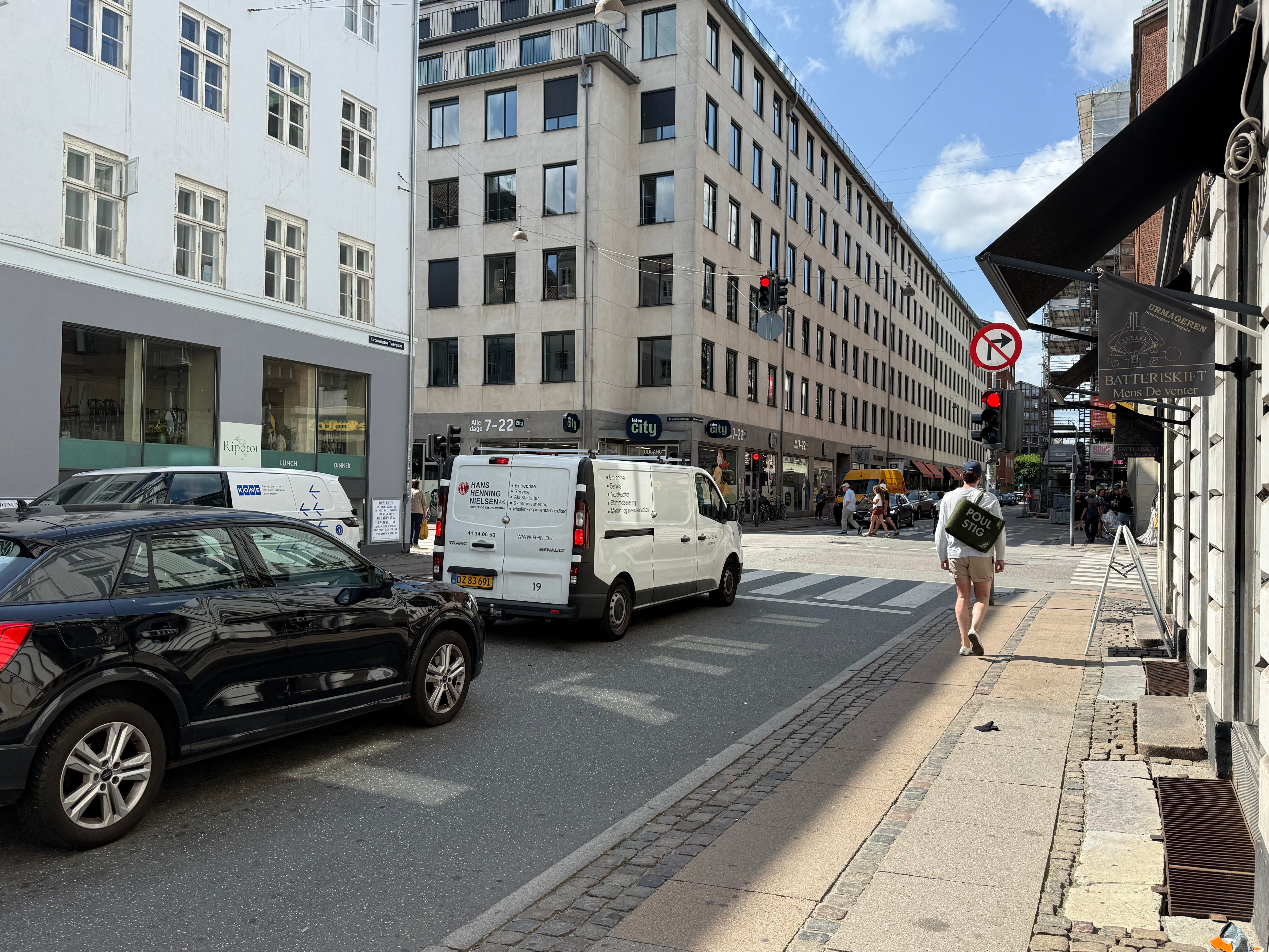 En bil og en van er parkeret og står i lyskryds. En person går på fortov med en rygsæk. En stor bygning med teksten City på