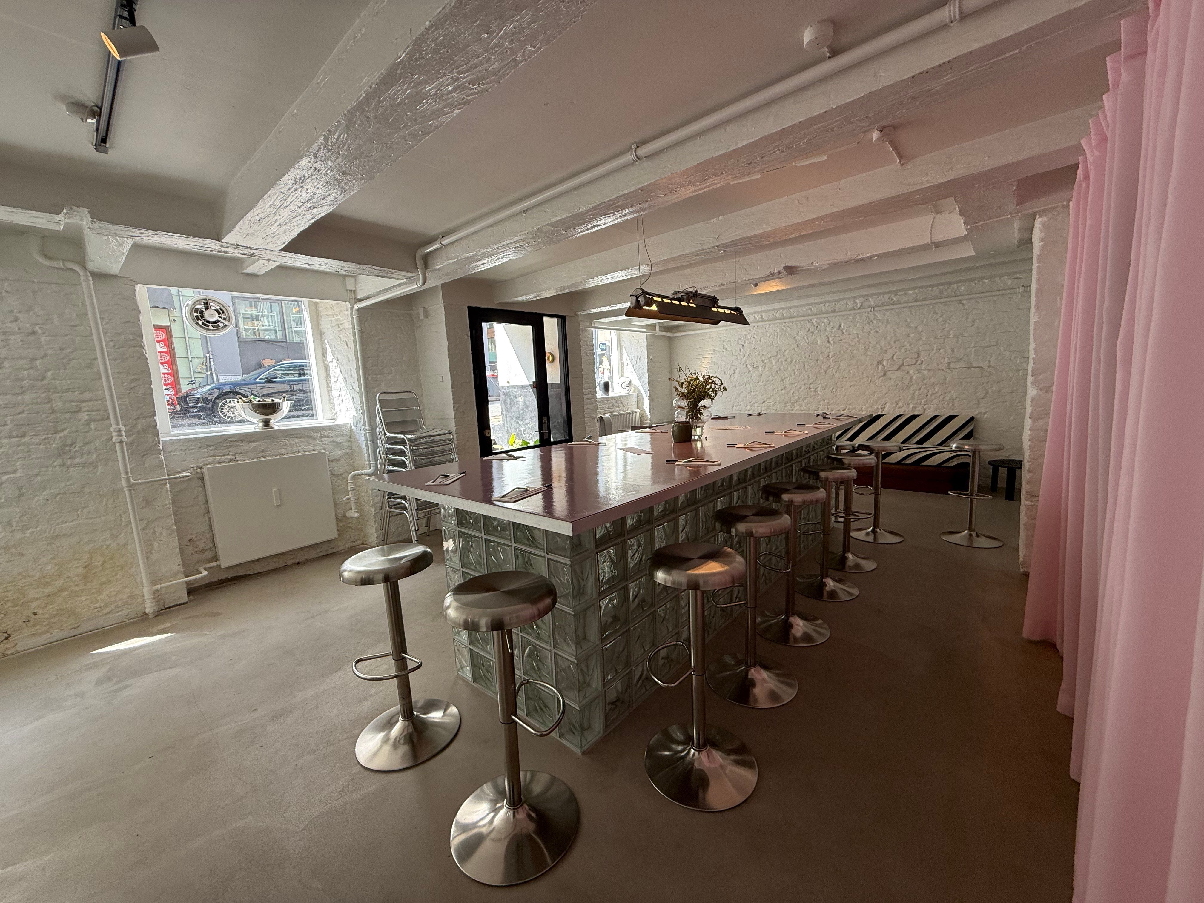 Højloftet lokale med glasbar, stolpebord, stole og tæppe på gulvet, samt udendørs stolpebord og stole