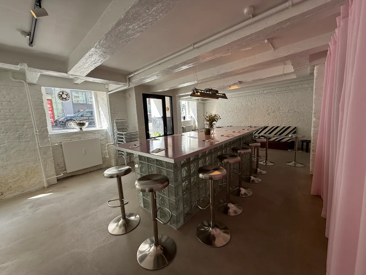 Højloftet lokale med glasbar, stolpebord, stole og tæppe på gulvet, samt udendørs stolpebord og stole