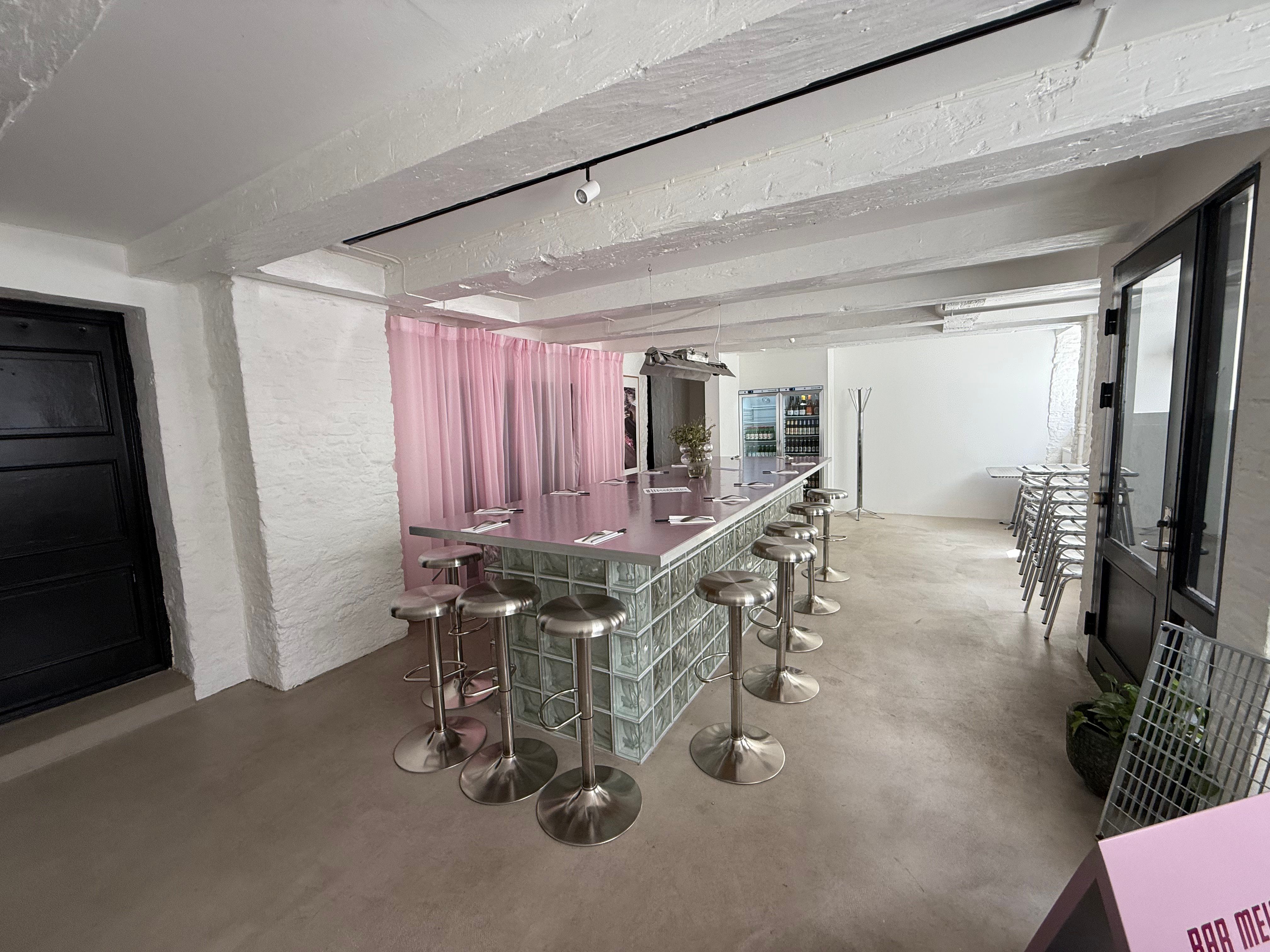 Langt, tomt køkken med højt loft og hvide vægge, og to store længe borde med stolpeforstærkning samt stole med metalfødder