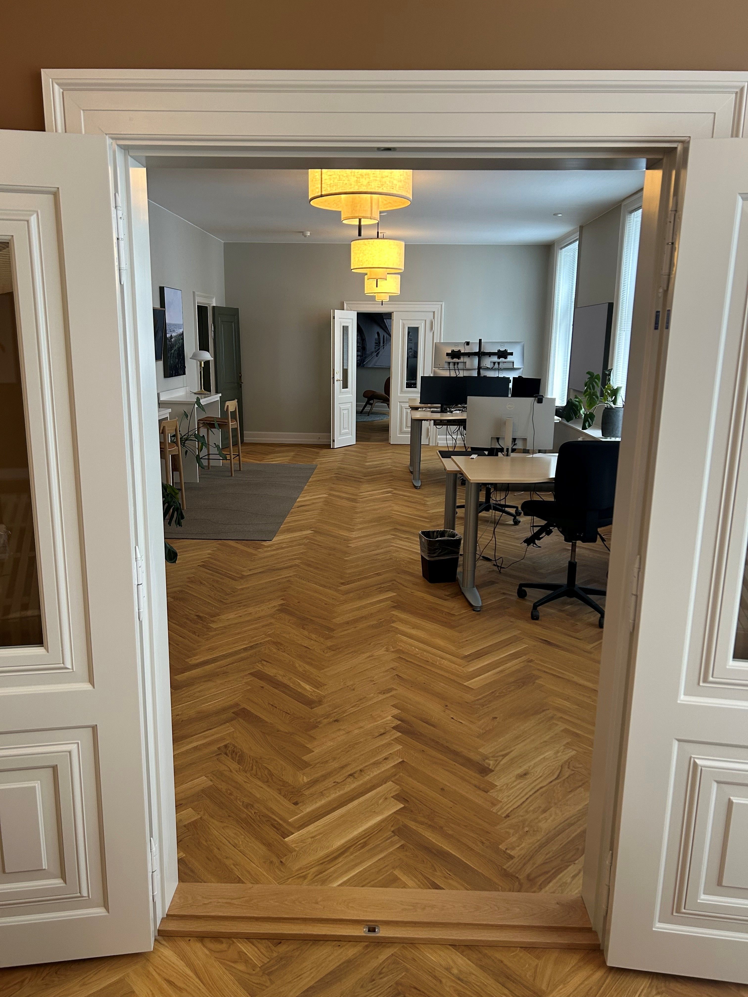 Åben dør til et kontorlokal med lyse gulve og lysekroner. Værelset er indrettet med skriveborde, stole og plante i hjørnet.