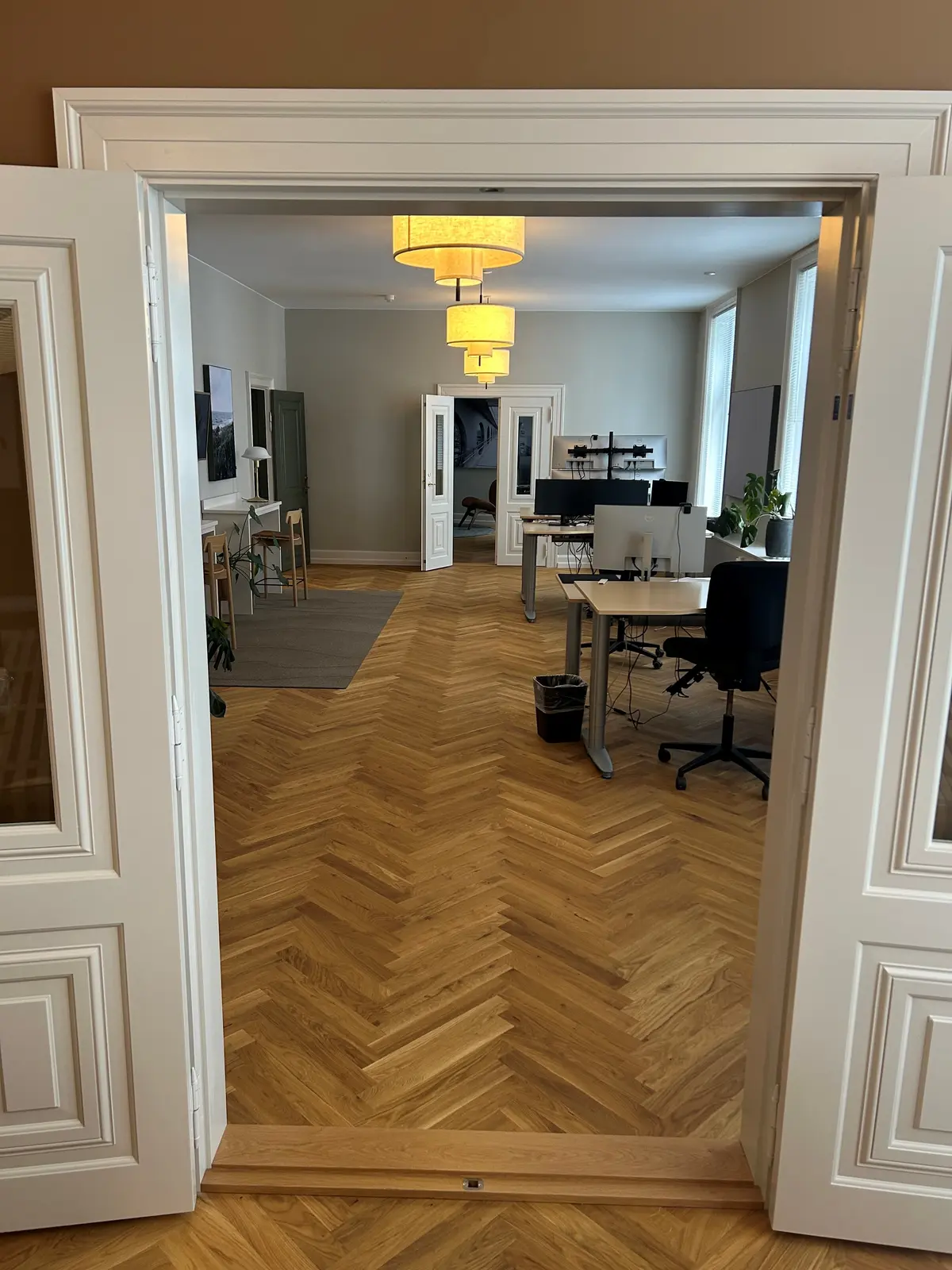 Åben dør til et kontorlokal med lyse gulve og lysekroner. Værelset er indrettet med skriveborde, stole og plante i hjørnet.