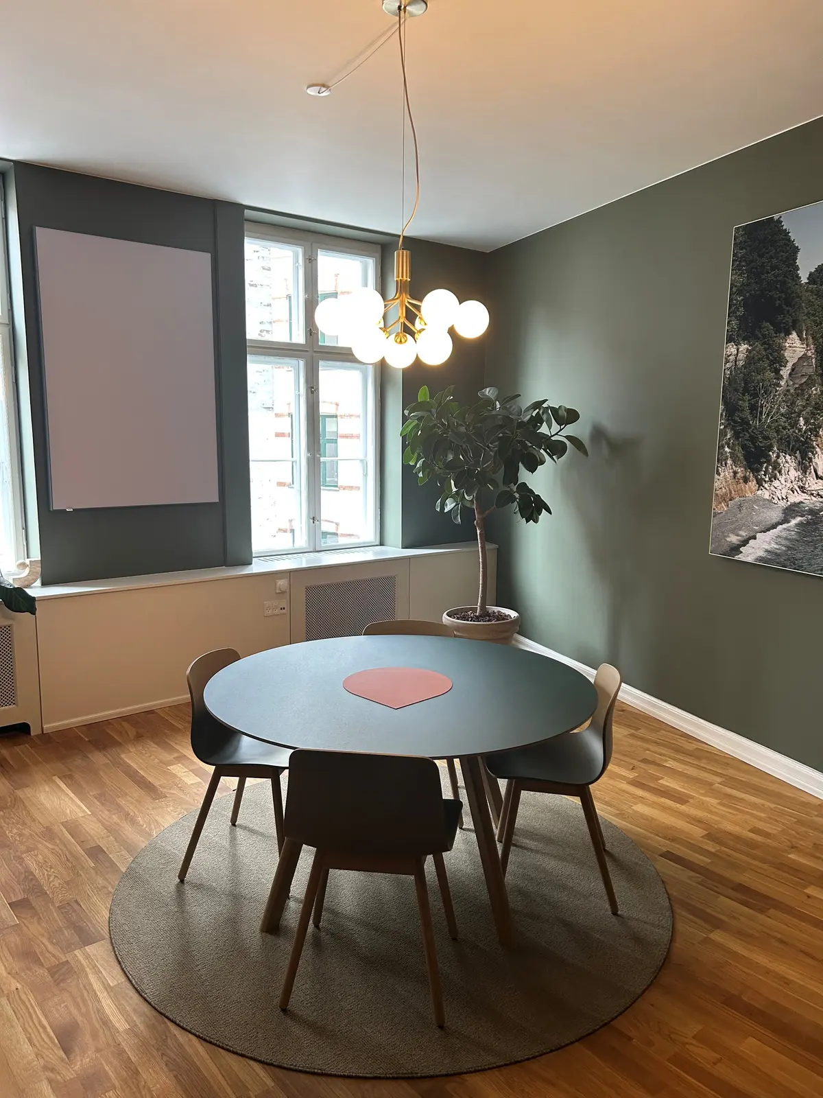 Rundt spisebord med fire stole omkring og en stor plante i hjørnet med en lysekron med flere kugler over bordet