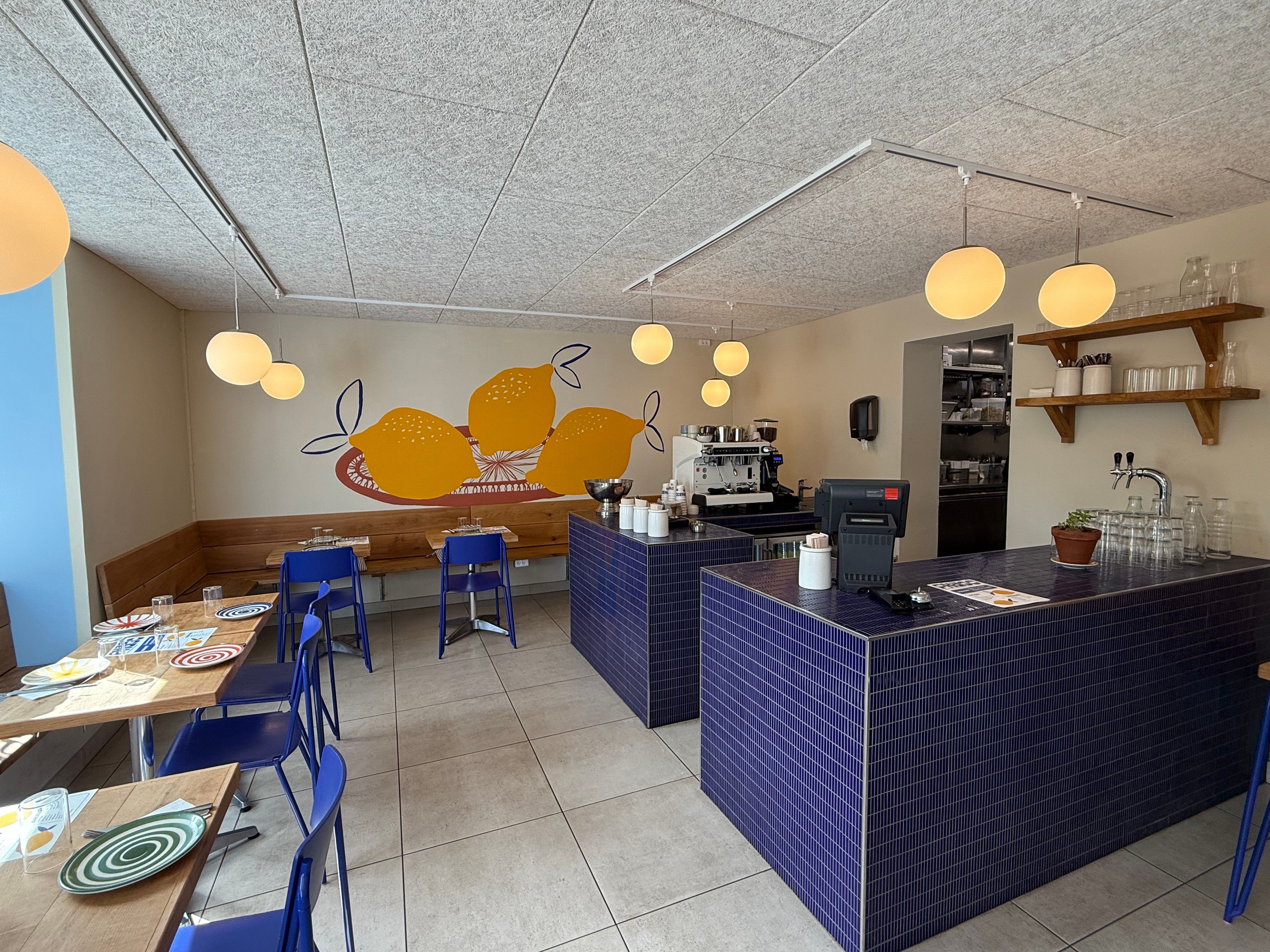 Inde i en café med borde, stole og lamper samt udsigt til en terrasse med planter og glas