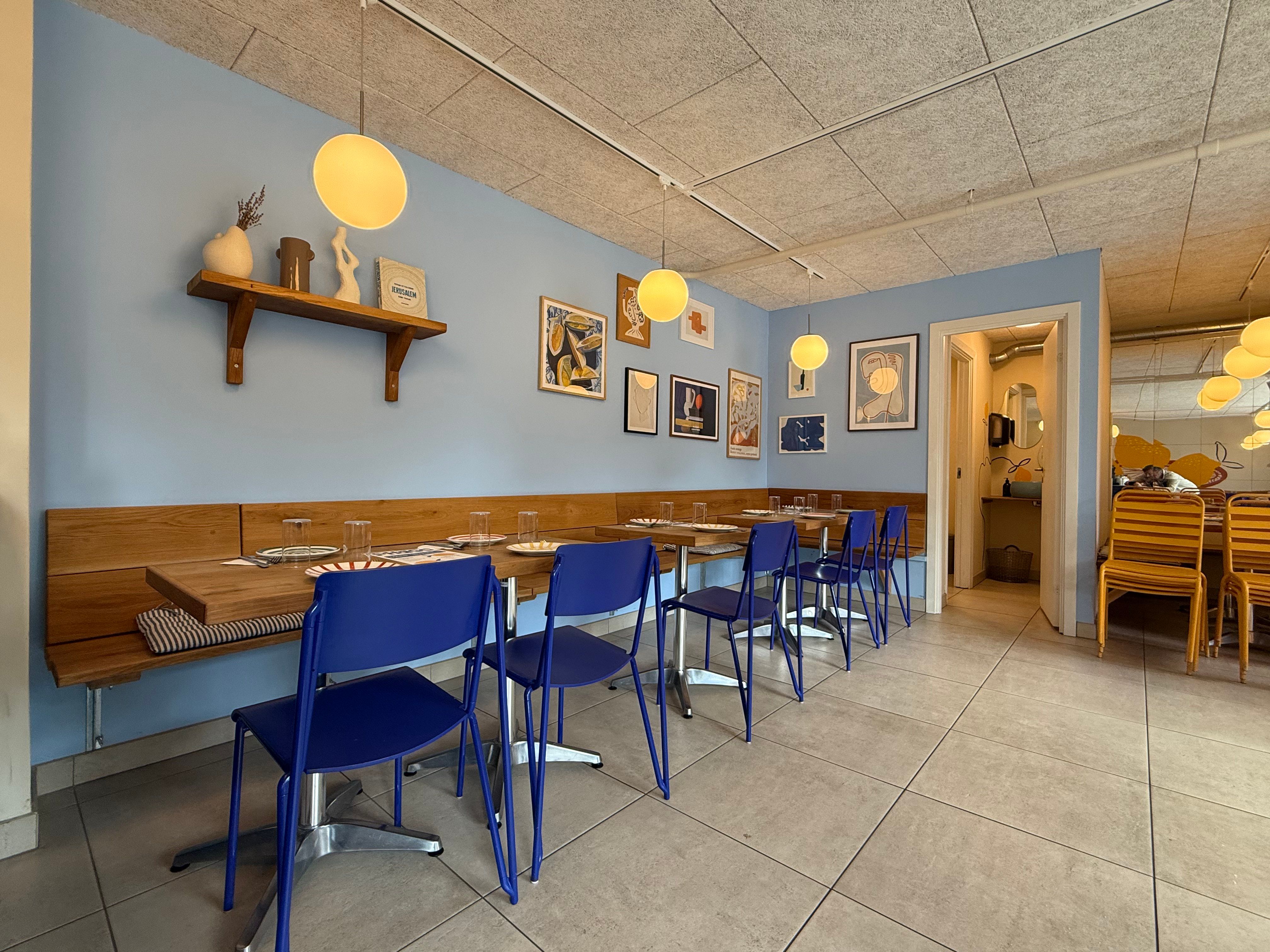 Lyst og rummelig spisesal med blå stole og lyse loftslamper. Bordplader med tallerkener og glas samt hylde med kunstværker.