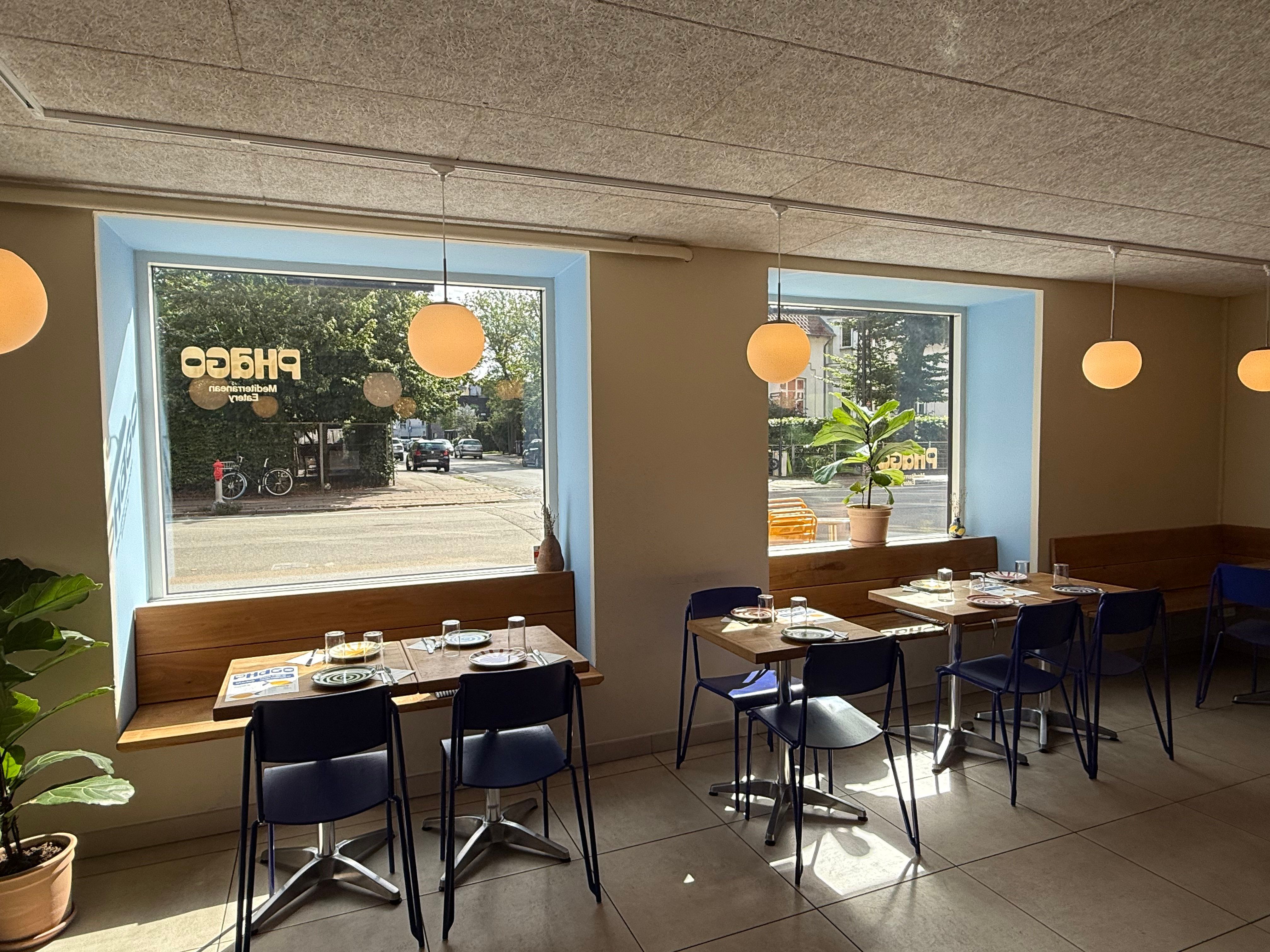 Et lille, hyggeligt café med bord og stole og en plante foran vinduet og udsigt til gaden med biler og cykler