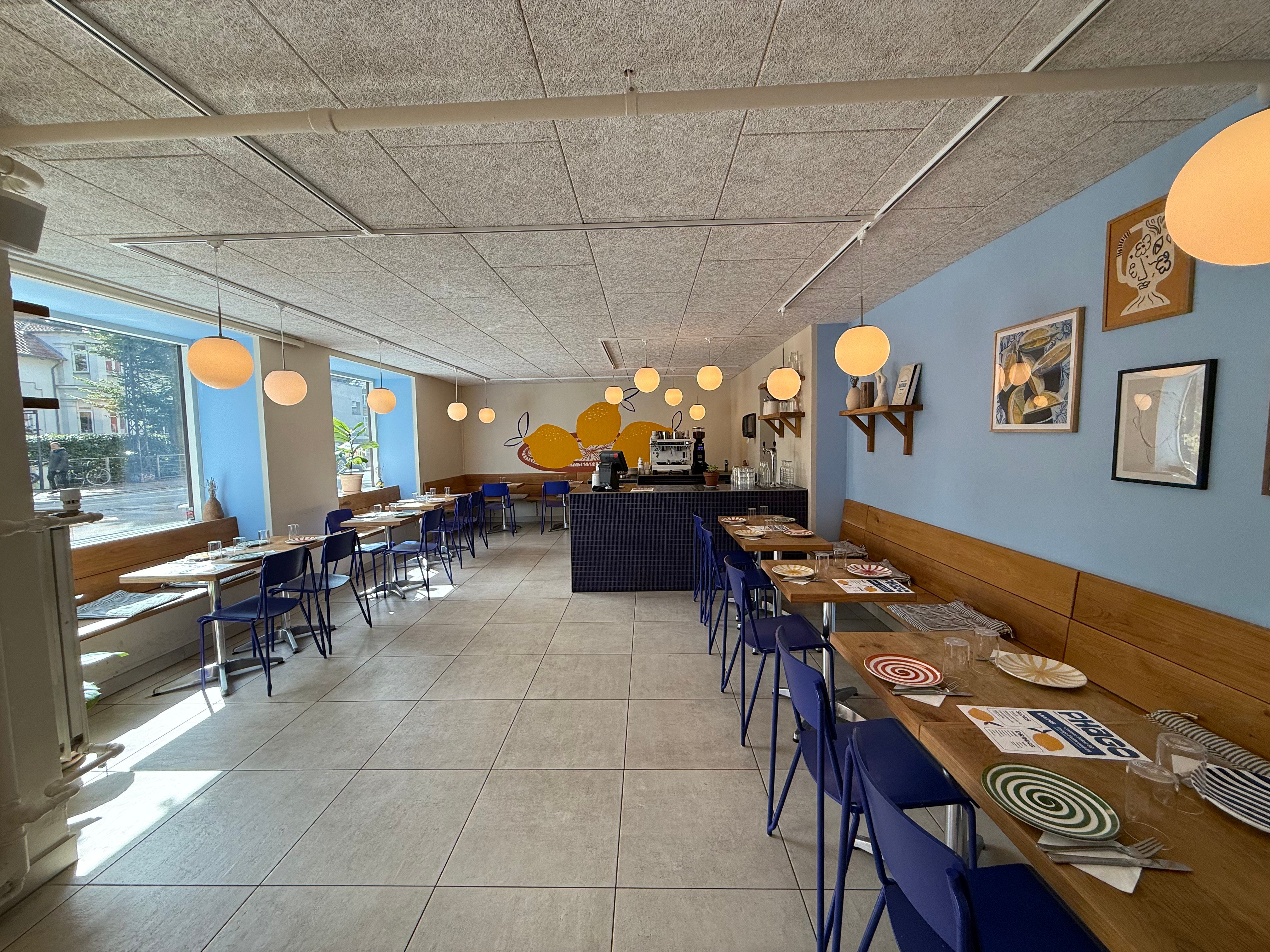 En tom restaurant med lyse blå stole og borde, lysekroner og malerier på væggen, og udsigt til bygninger og planter udenfor