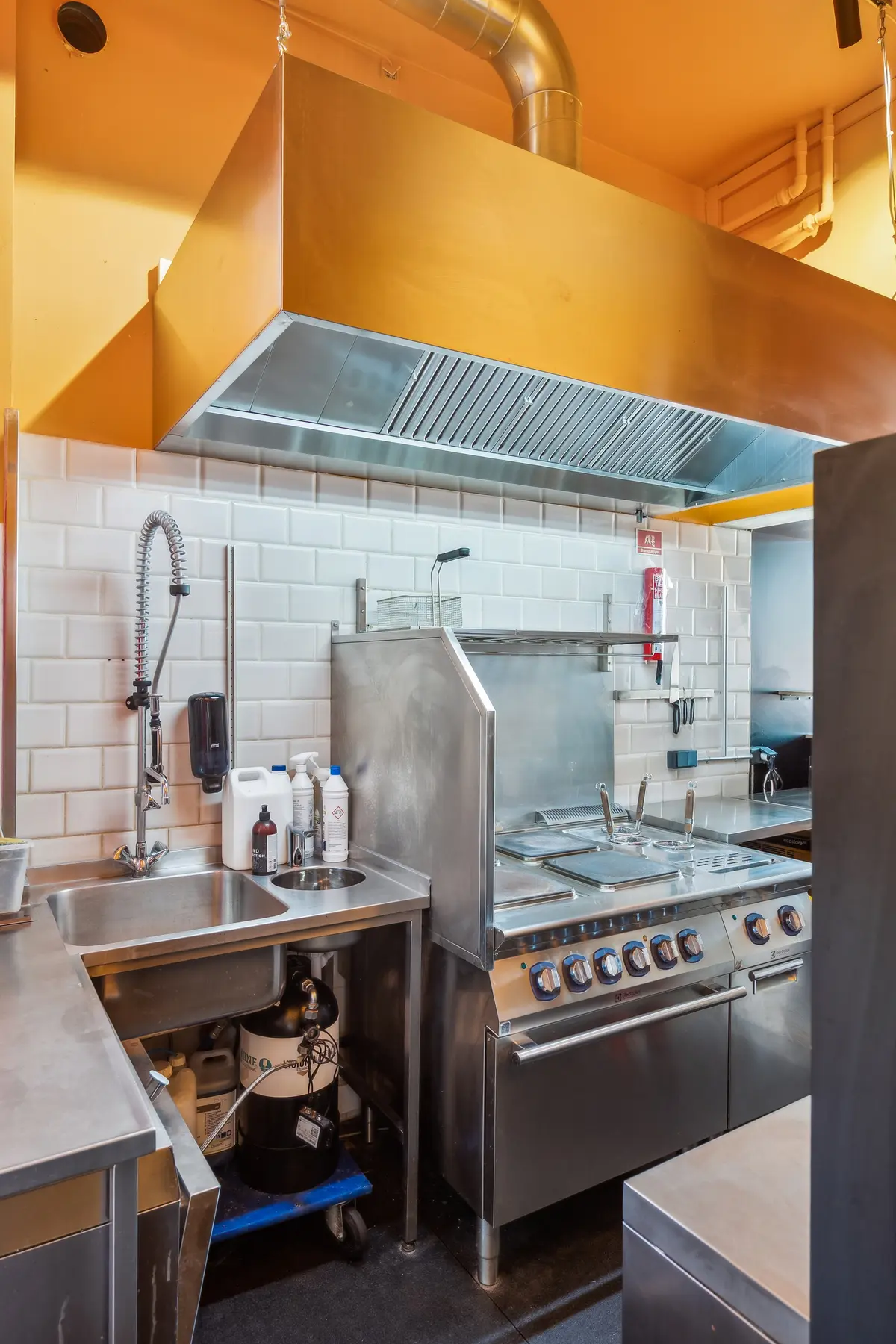 En moderne kommerciel køkken med stålapparater og en stor ventileringshatte over ovnen og komfuret, samt et vaskebælte med