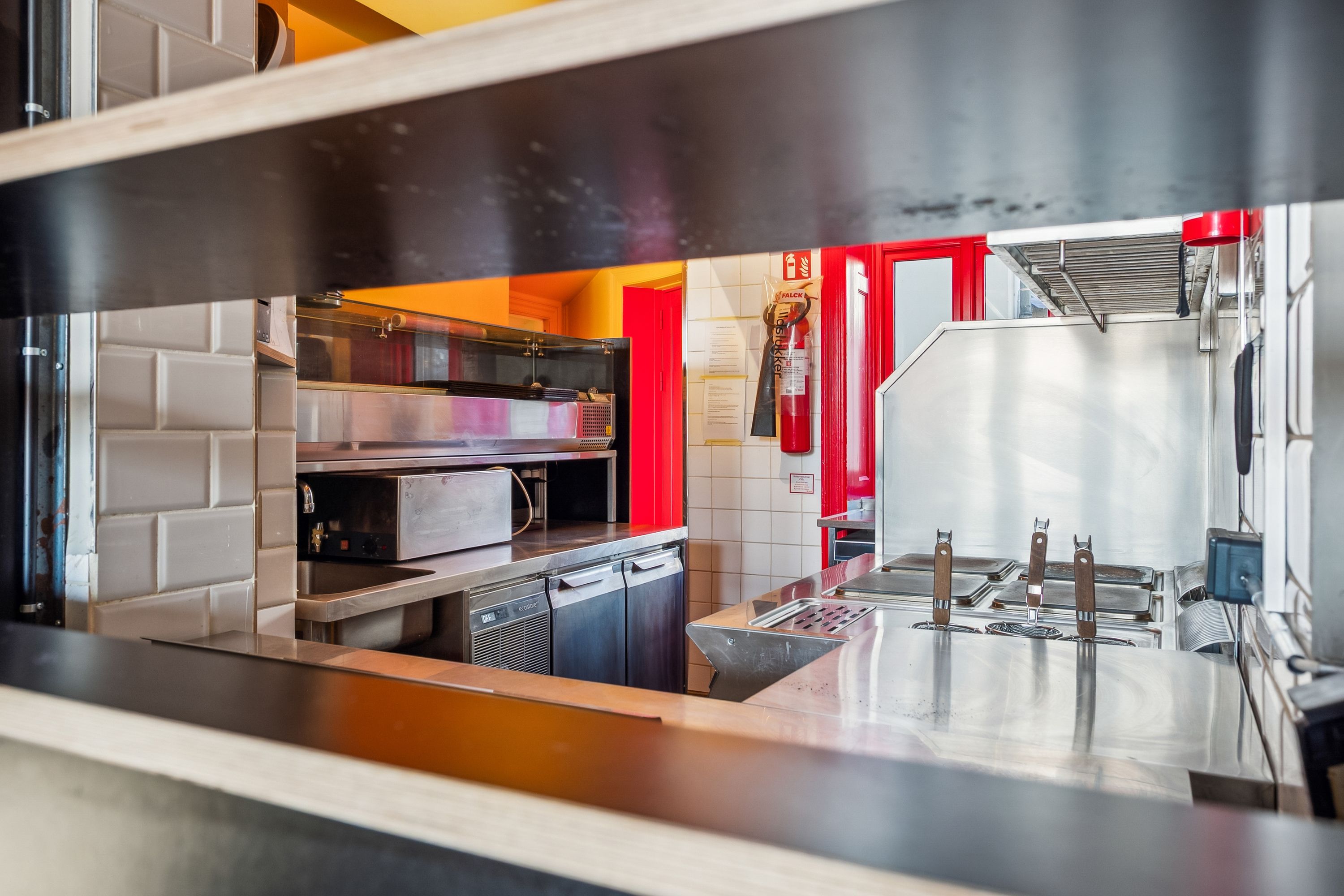 Rustfrit stål køkken med kogeplader, ovne, skabe og en ildslukker væg. Refleksioner og skygger vises på overfladen af