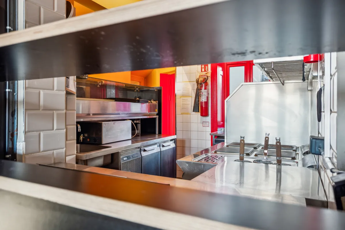Rustfrit stål køkken med kogeplader, ovne, skabe og en ildslukker væg. Refleksioner og skygger vises på overfladen af