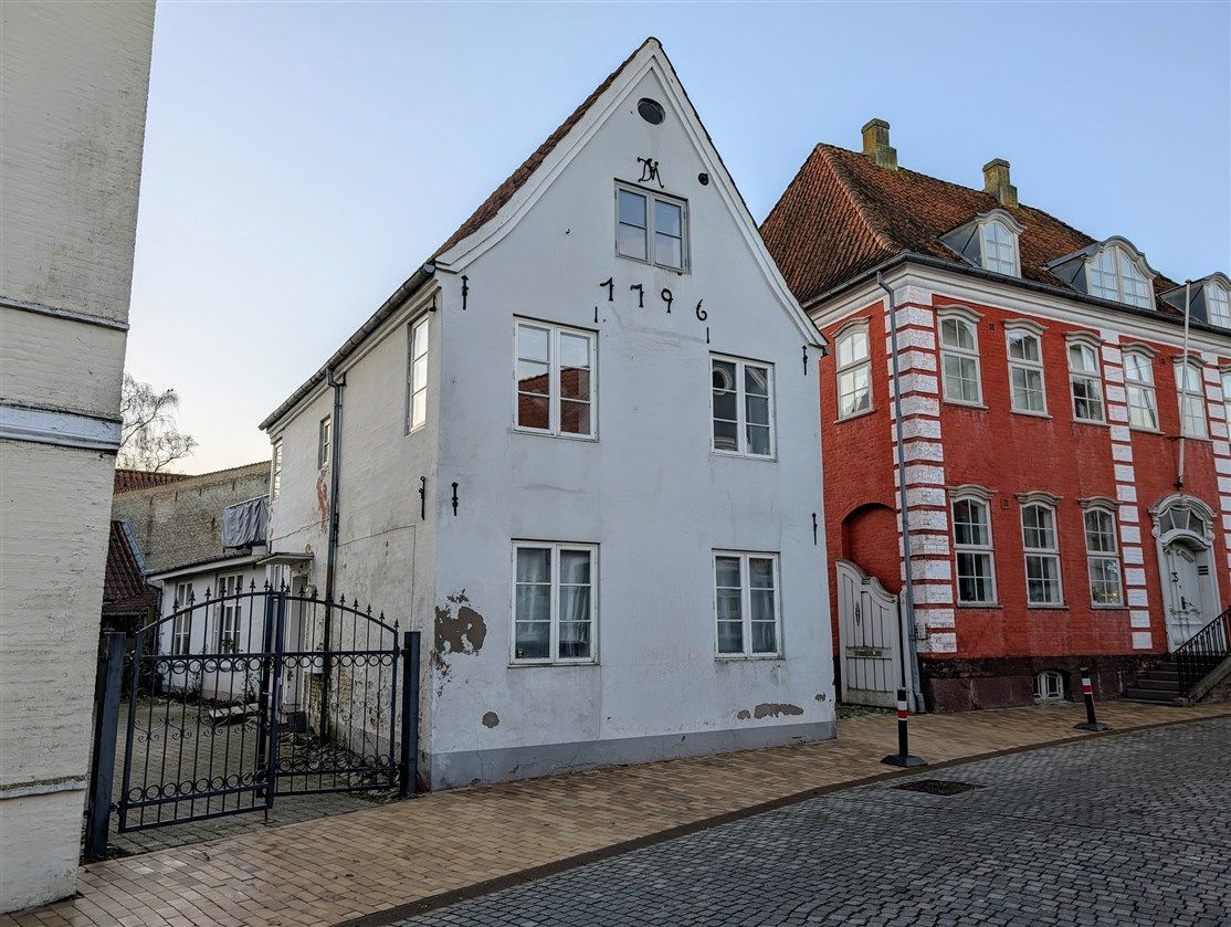 Hjørnebygning med røde mursten og rødt tag i Aabenraa, hvid bygning med nummer 1196 og skrånende tag