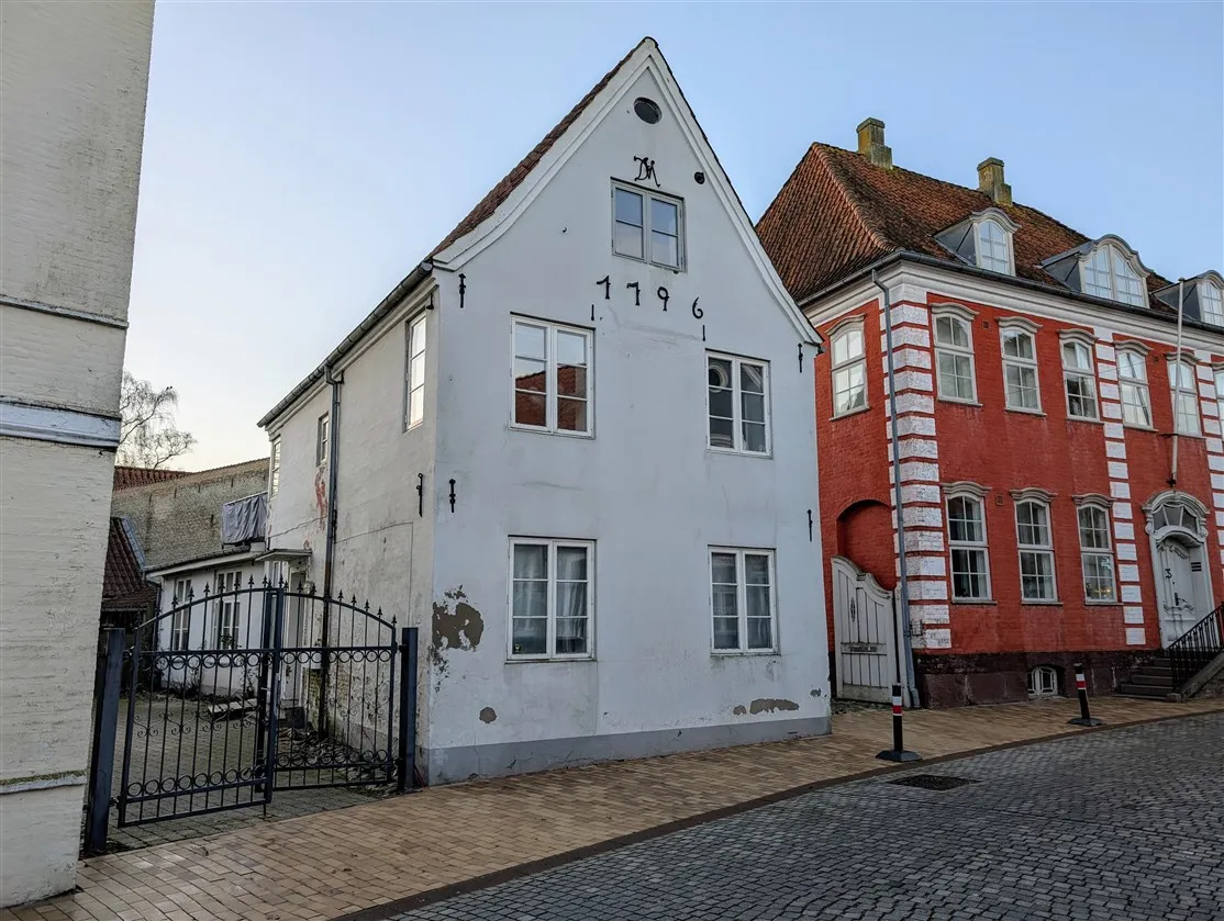 Hjørnebygning med røde mursten og rødt tag i Aabenraa, hvid bygning med nummer 1196 og skrånende tag