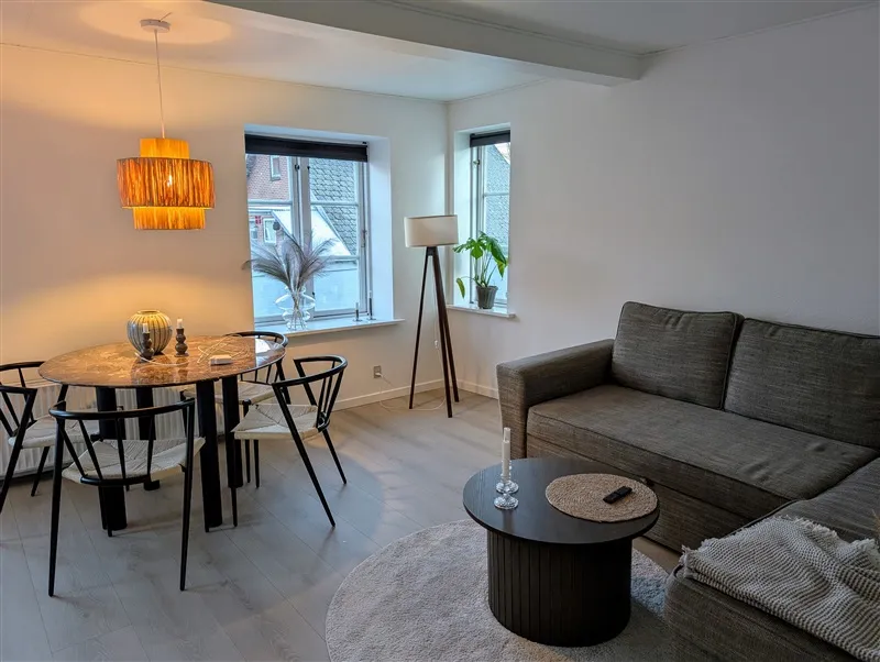 Lys stue med spisestue, sofa, runde bord, lampe og vindue med plante