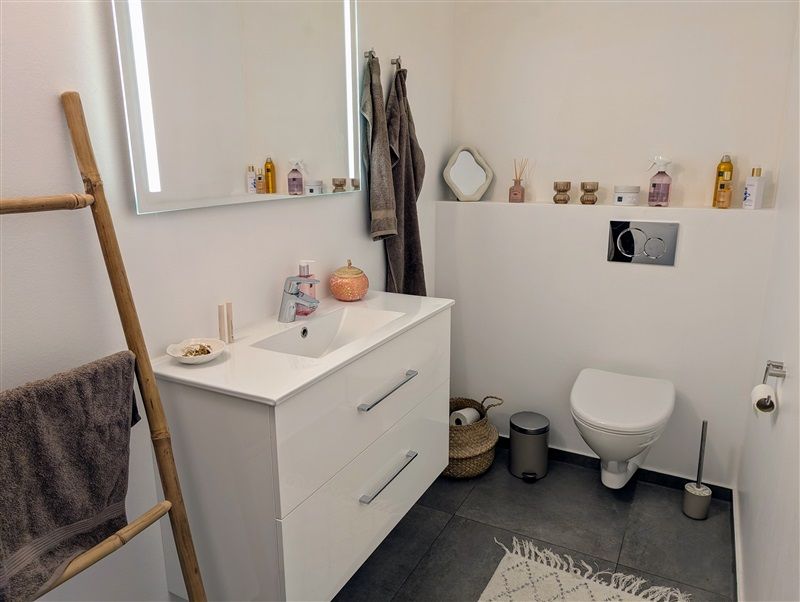 En moderne badeværelse med hvidt badeværelsesskab og undertøjshylde, hvidt toilet, gulvtæppe og et spejl med refleksion af