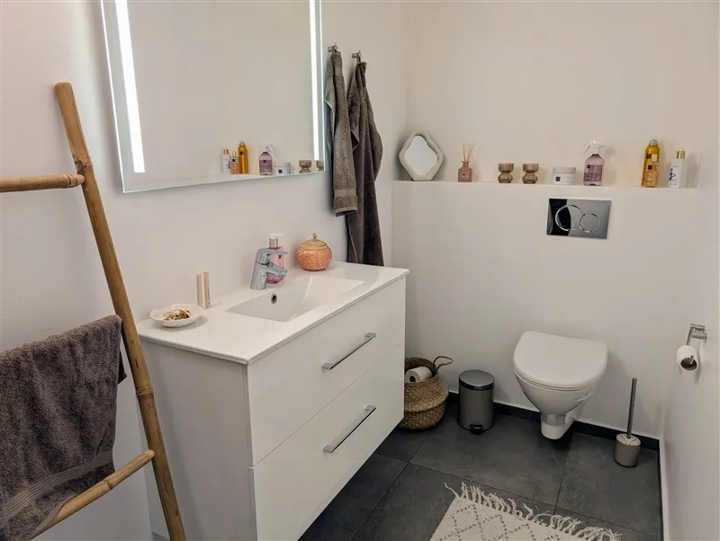 En moderne badeværelse med hvidt badeværelsesskab og undertøjshylde, hvidt toilet, gulvtæppe og et spejl med refleksion af