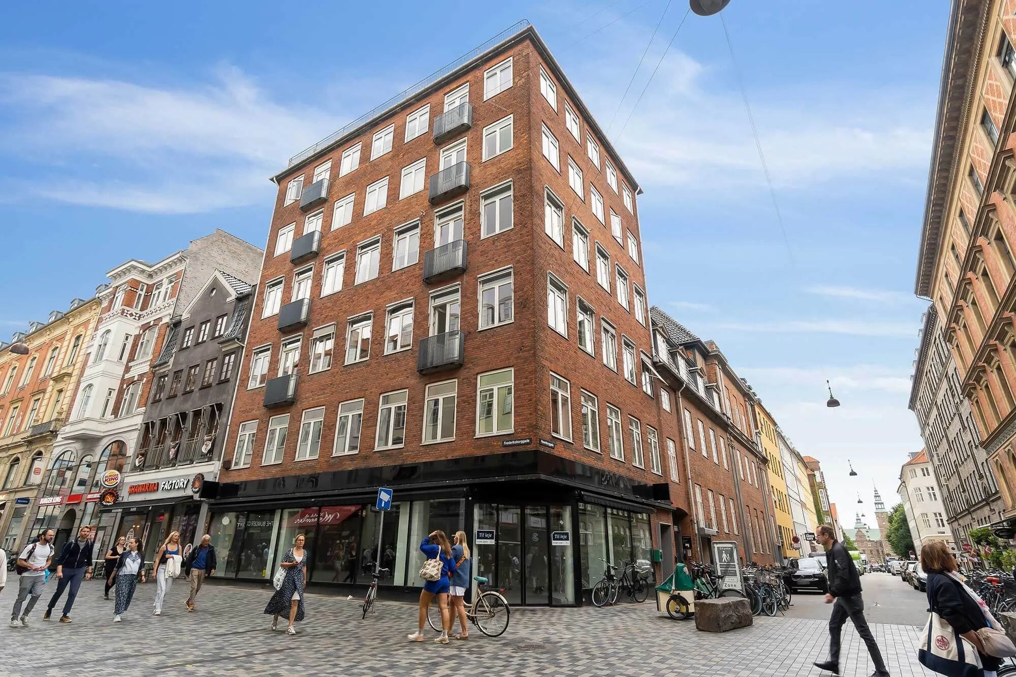 Folk går på en gadehjørne butik med store glasvinduer og facader mod gaden Frederiksborggade, der er fuld af cykler og biler