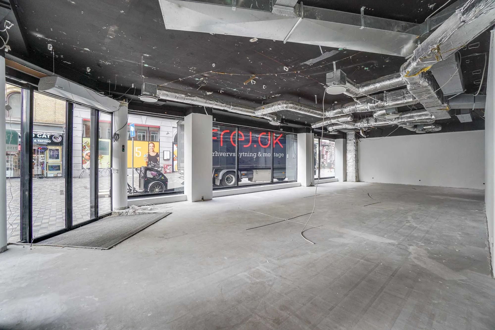 Frederiksborggade med søjler, glasdøre og plakater, tomme lokaler med kabelslange og ventiler, bus og skulpturer i baggrunden