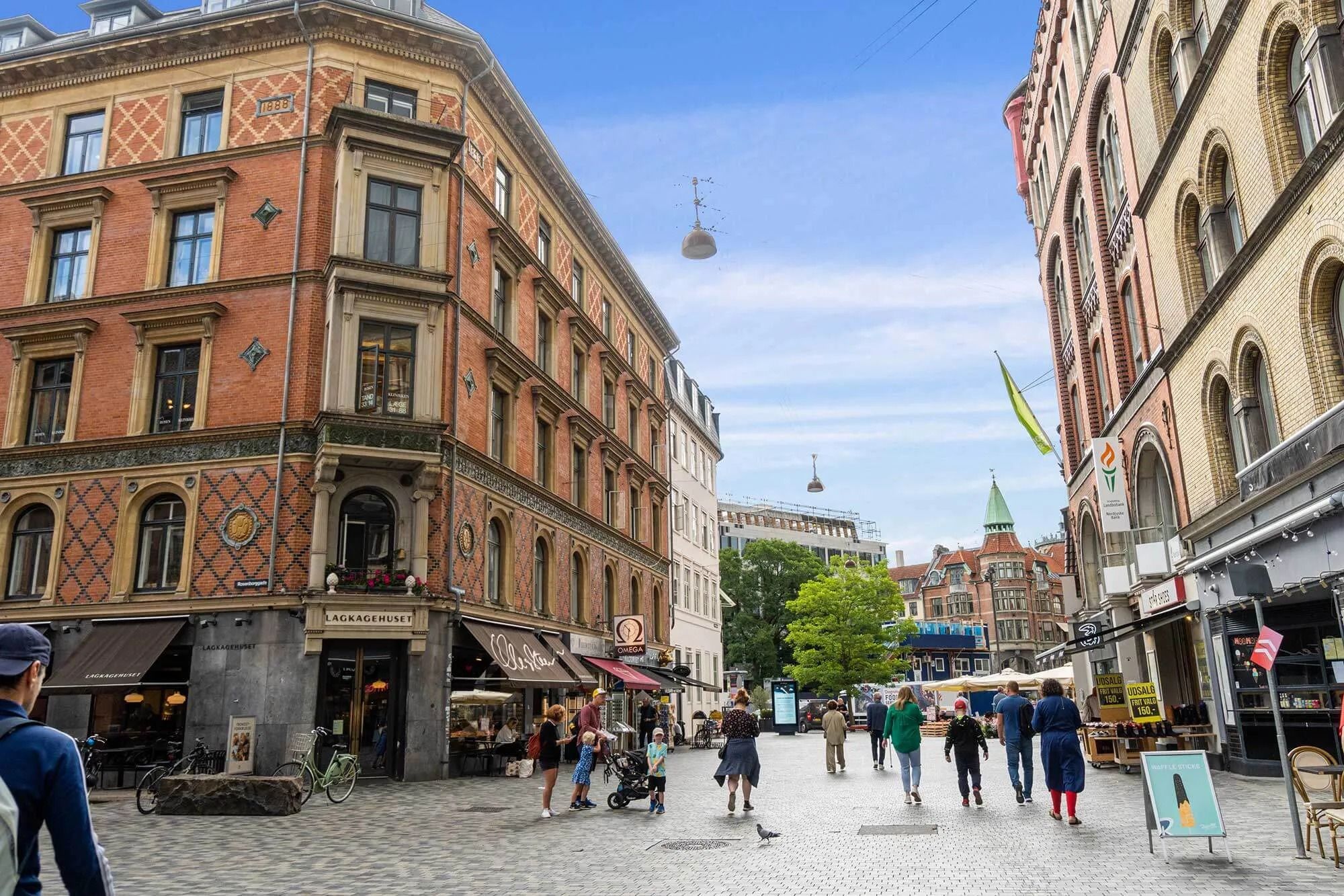 Hjørnebygning med flere etager og butikker, hvor folk går og står og gåkøre i Frederiksborggade, med en banner med teksten