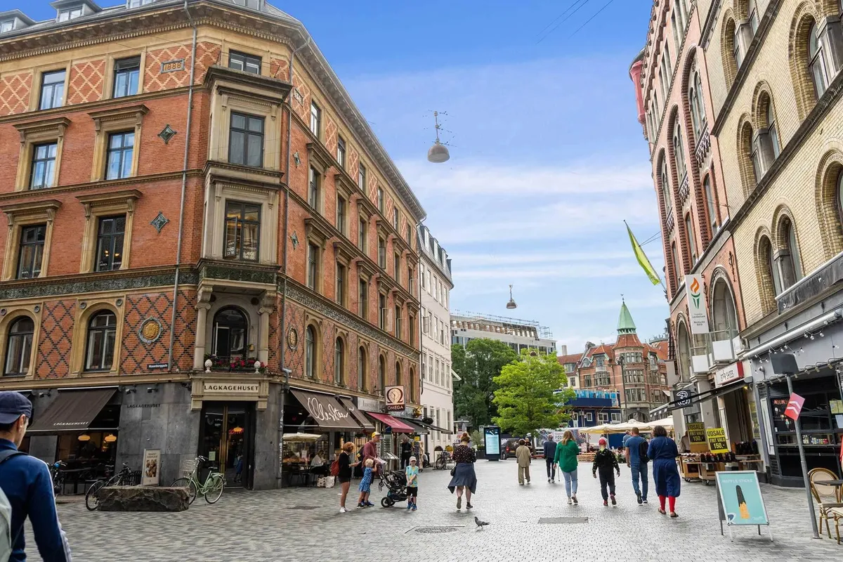 Hjørnebygning med flere etager og butikker, hvor folk går og står og gåkøre i Frederiksborggade, med en banner med teksten