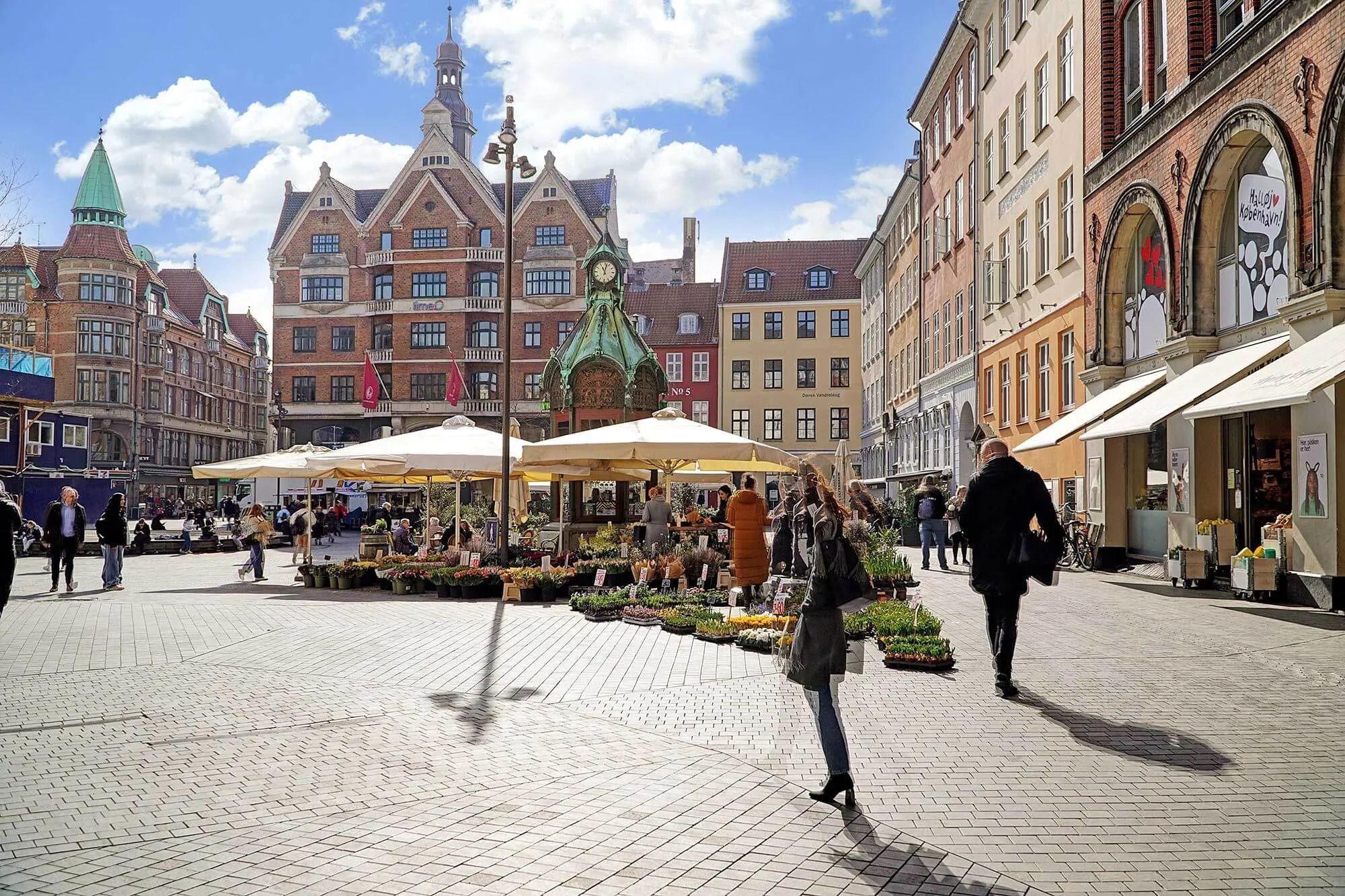 Lejet butik med facade ud mod torvet, folk går forbi, og der er mange blomster og planter i potter