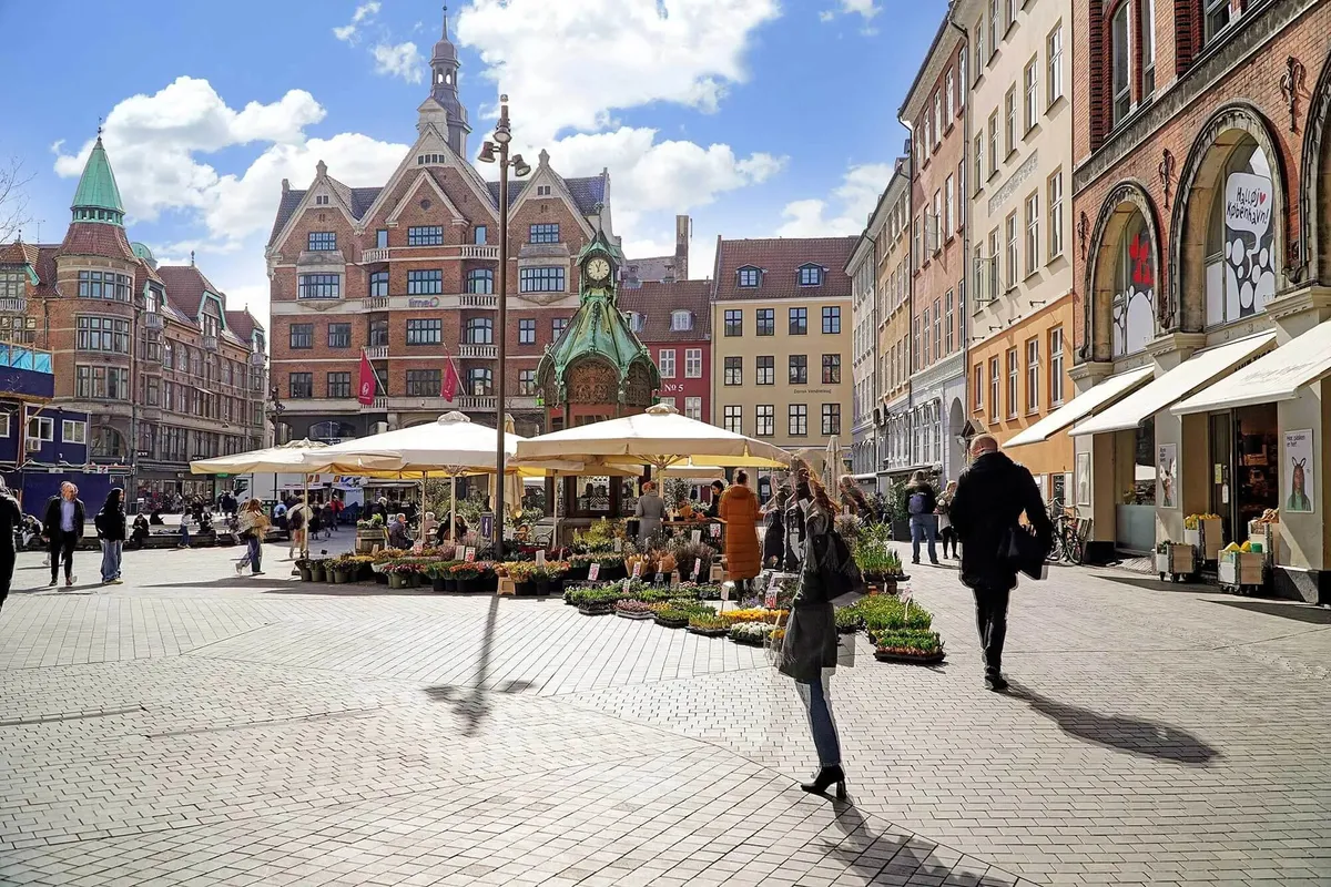 Lejet butik med facade ud mod torvet, folk går forbi, og der er mange blomster og planter i potter