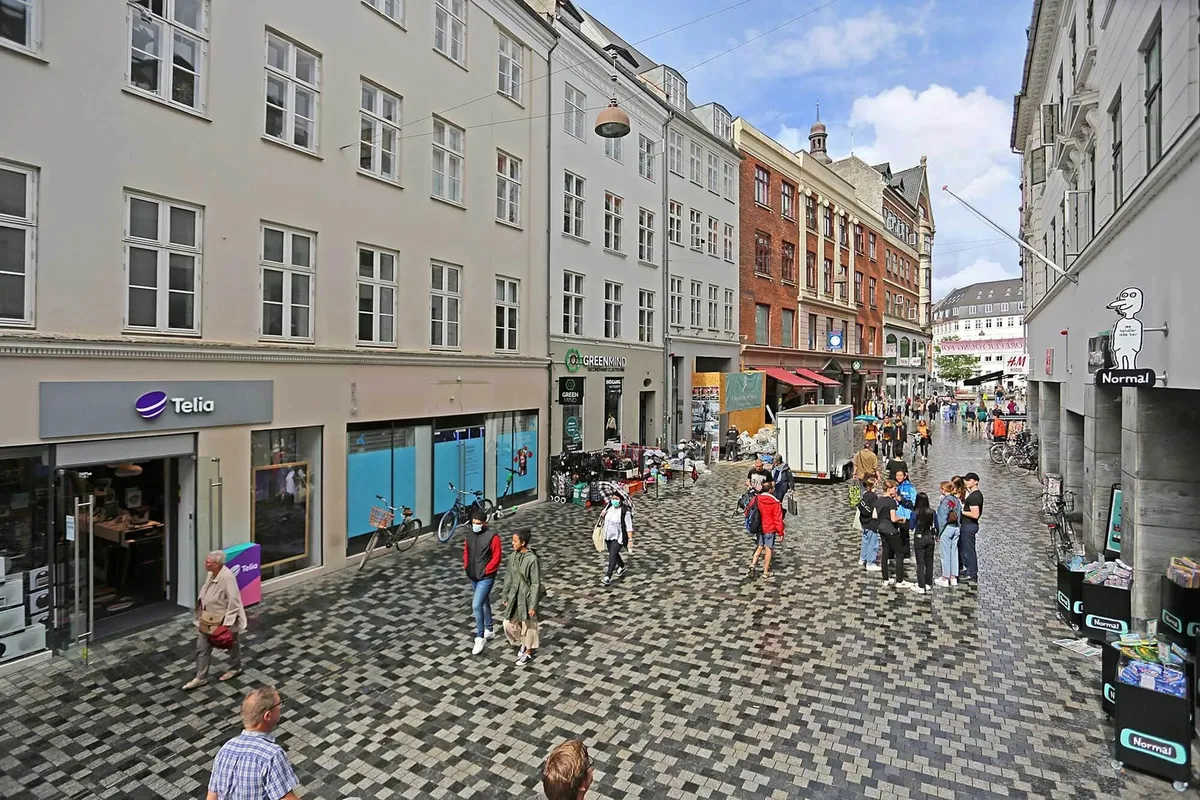 Mennesker vandrer rundt på Købmagergade, forbi butikker og en Telia-reklame, mens en buske cykler forbi