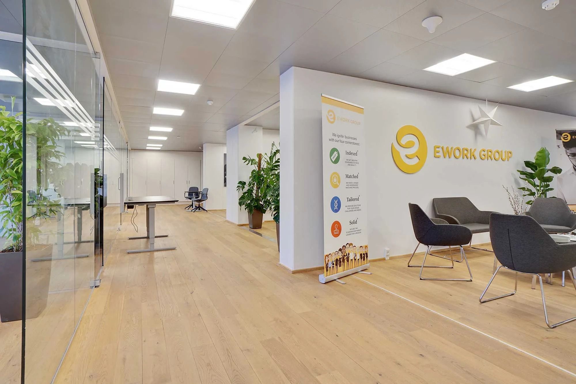 Kontor med lyse lofter og store glasruder viser EWORK GROUP logo, rekreative stole og lysegulve samt flere planter