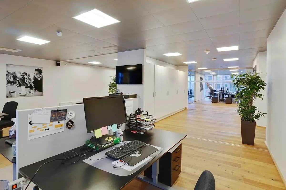 Kontorrum med trægulv, højt gulvloft og lyse loftslamper, møbler, to monitorer og et skrivebord med en tastatur, en mus og