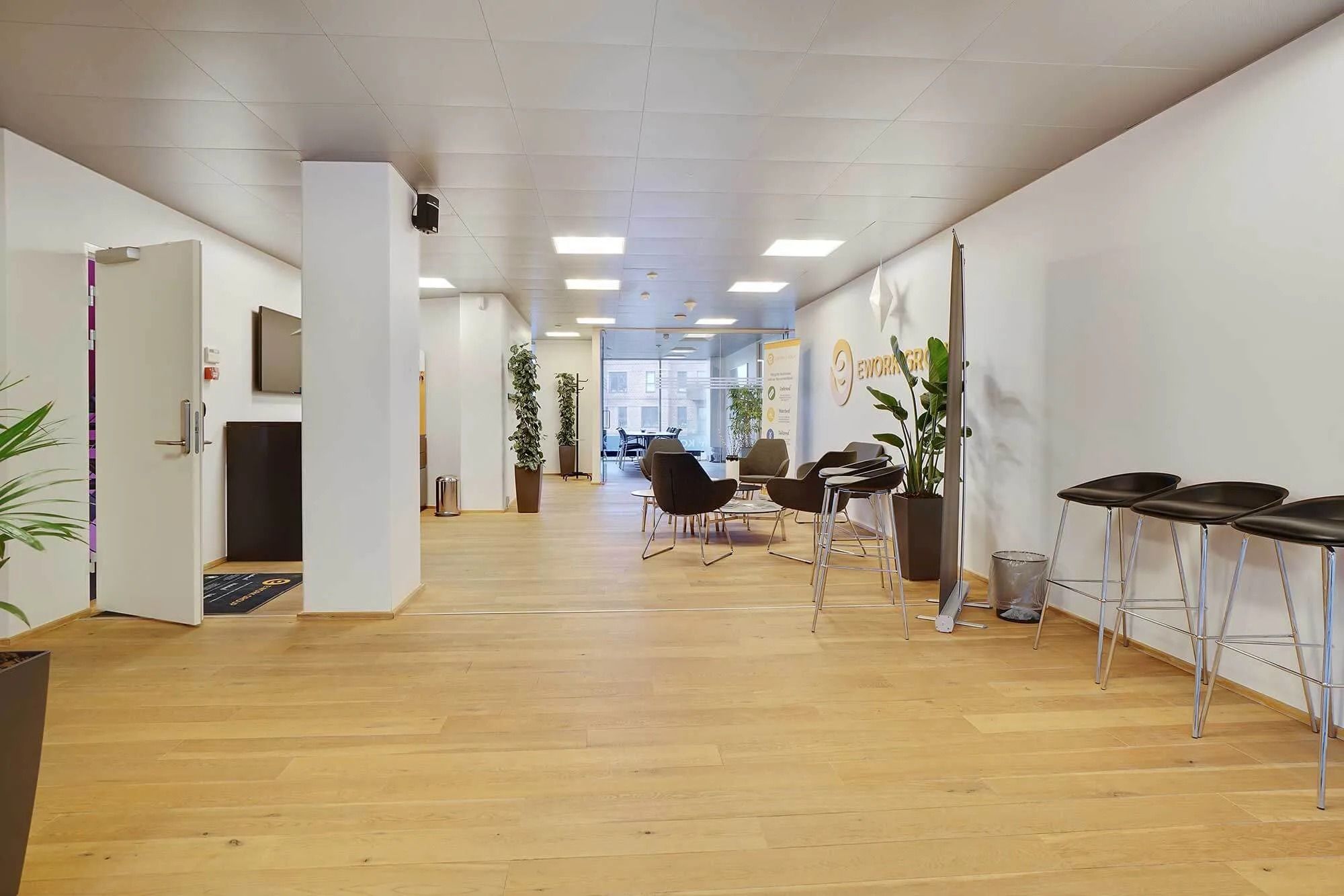 Kontorområde med højt loft, lysegul gulvbelægning og lysepunkter samt møbler og planter i rummet