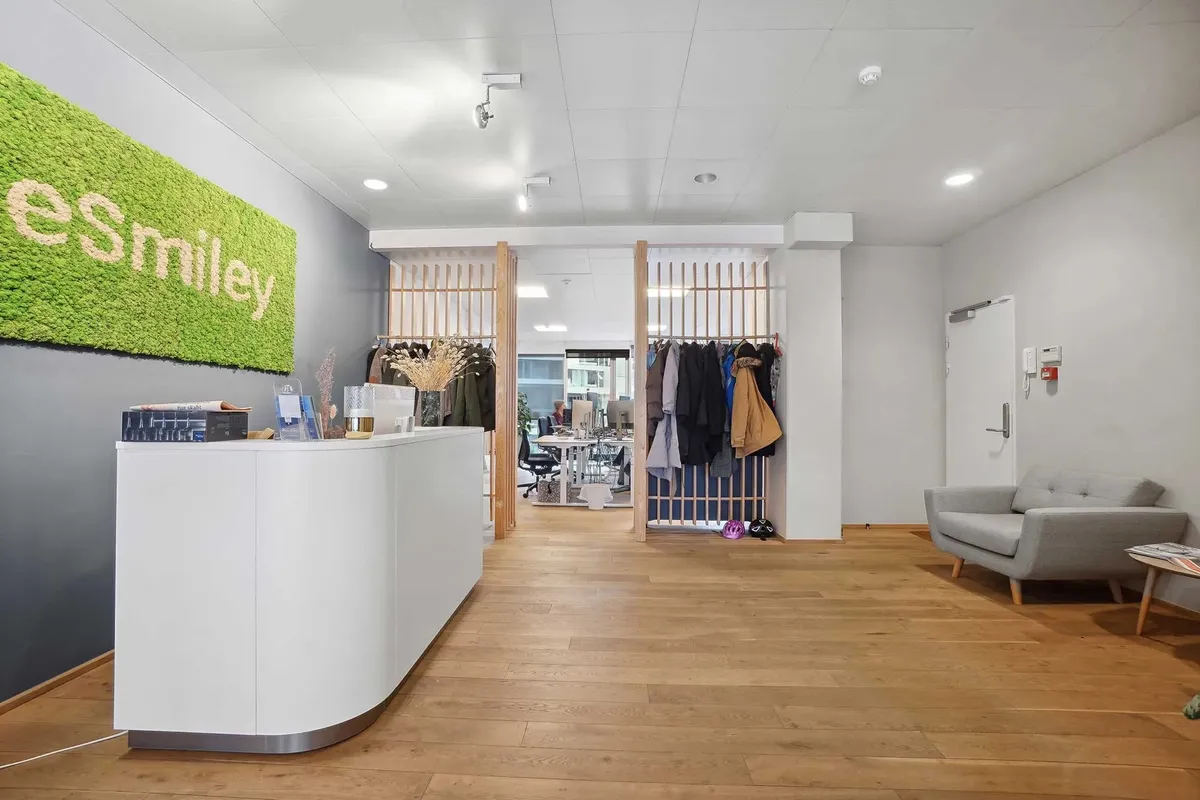 Kontor med en hvid receptionsbord, sofa og trægulv med en stor væg med grøn mos og teksten Esmiley