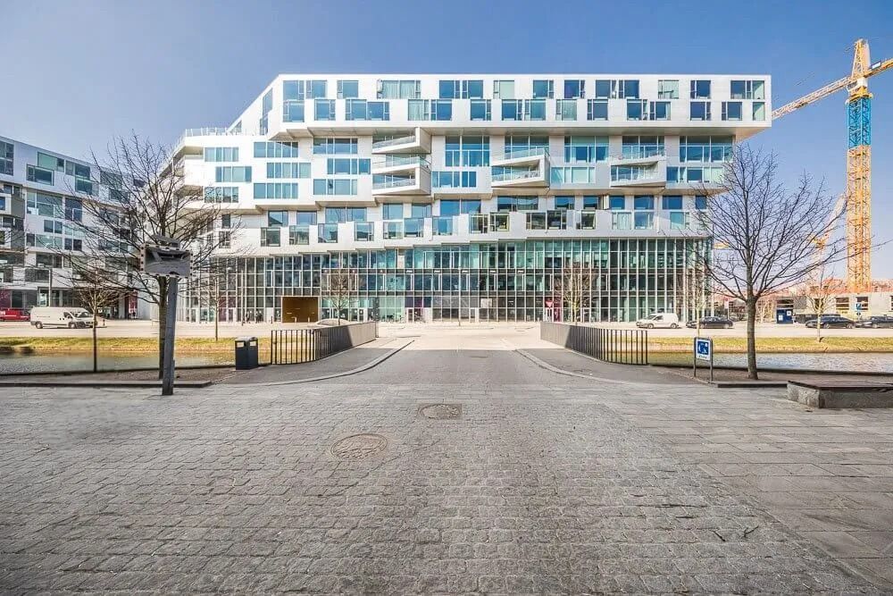 Moderne bygning med glasvinduer og parkeringsplads med biler og træer i forgrunden og kran i baggrunden