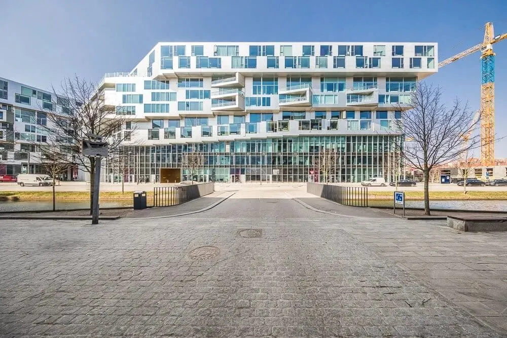 Moderne bygning med glasvinduer og parkeringsplads med biler og træer i forgrunden og kran i baggrunden