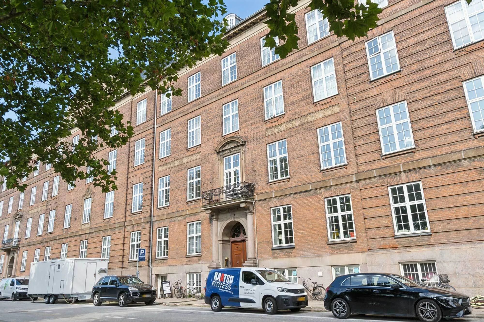 Kontorbygning med røde mursten og flere køretøjer parkeret udenfor, samt en trævogn og cykler