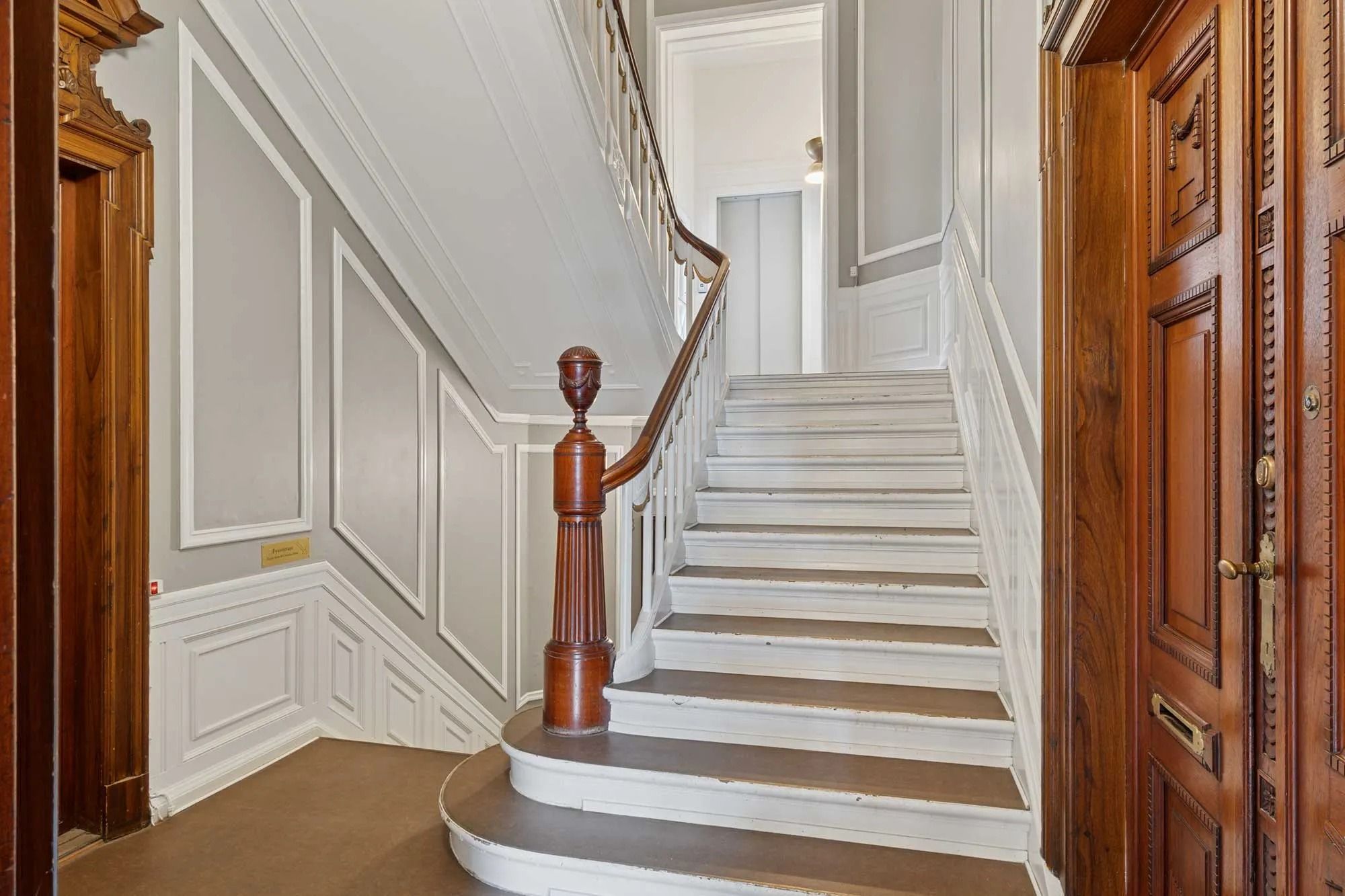 En elegant indgangsparti med høje, hvide vægge, træpaneler og en stor, slank trappe med lyse trin og træhåndtag. En åben dør