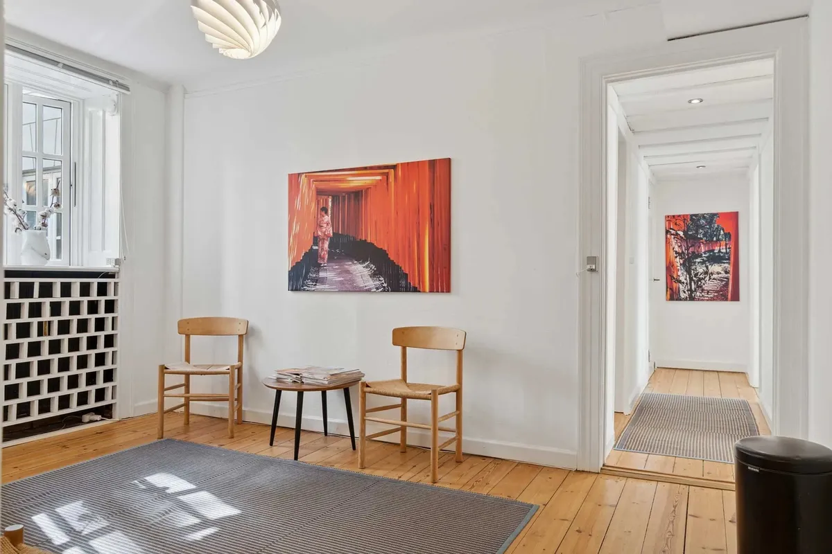 Værelse med hvide vægge, trægulv og en stor kunstværk. To stole og et bord med aviser. Værelse med gulvtæppe og åbent dør.