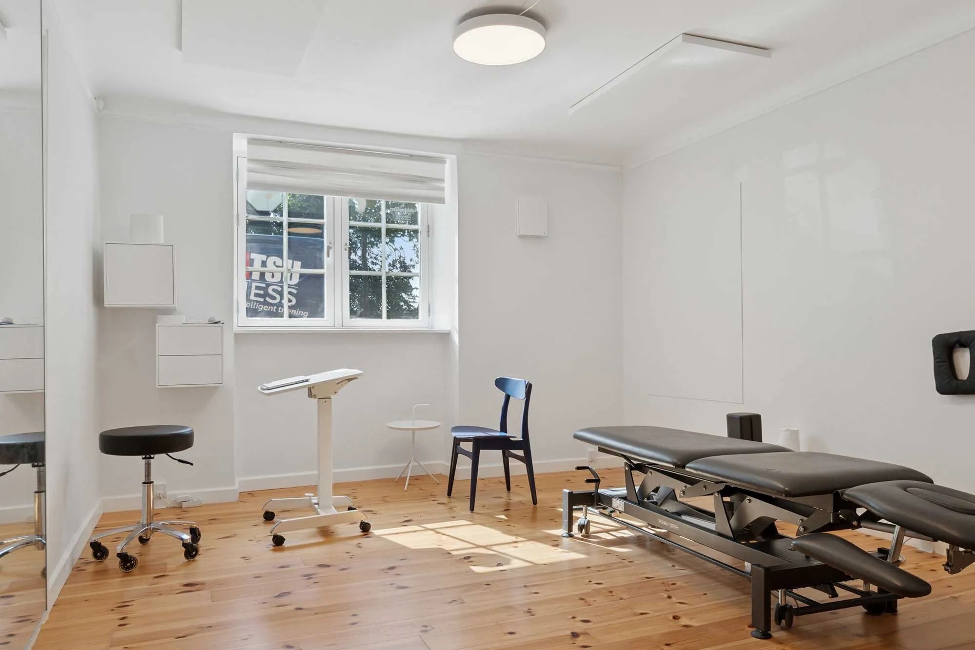 En lækker og pænt indrettet fysioterapirum med højloftede og hvide vægge og en stor spejl. Værelset har et stort vinduesglas