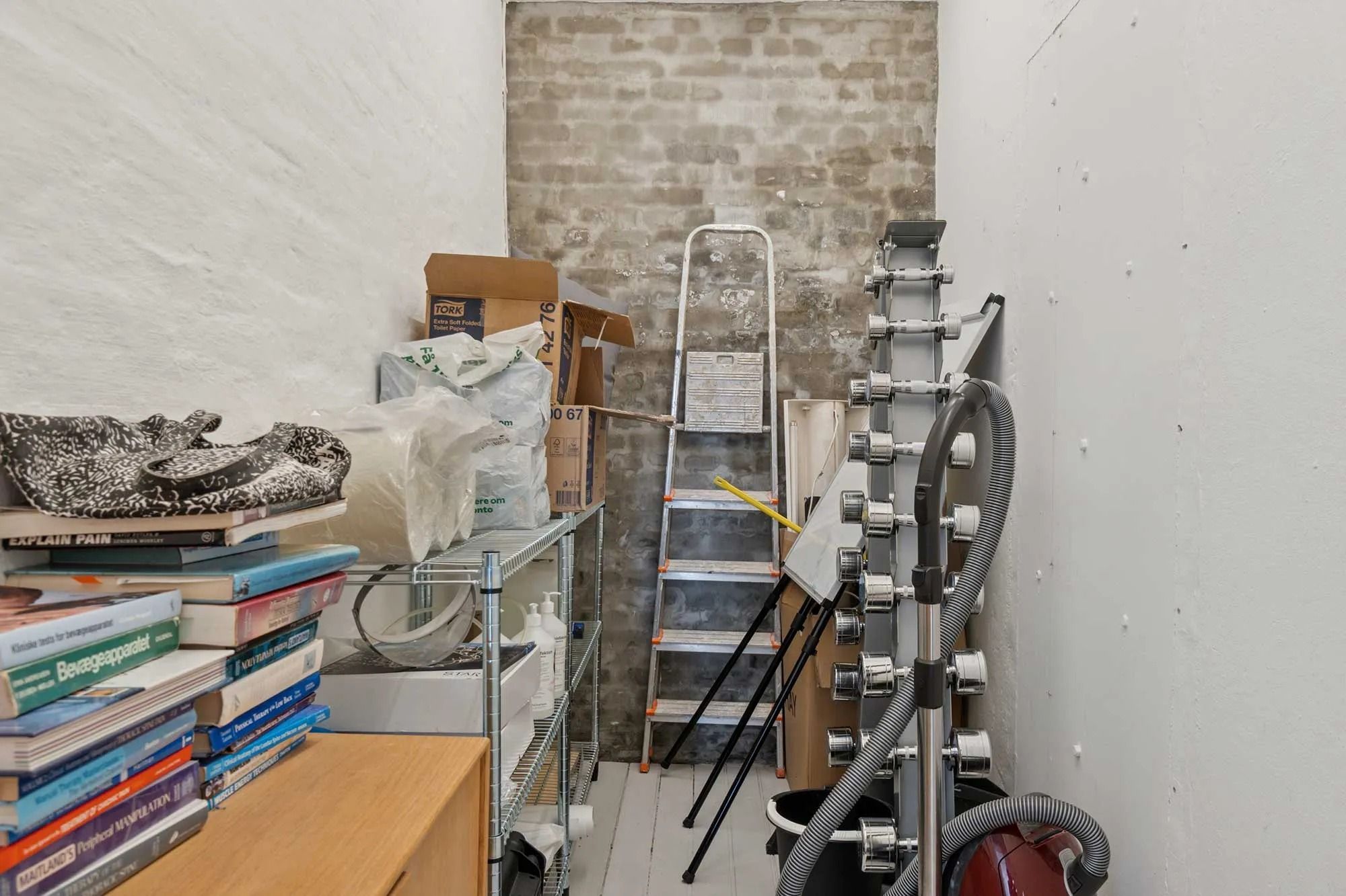 Lagerrum med bøger, sæbeflasker, en støvsuger, bokse og en stige mod en hvid og en gul mur