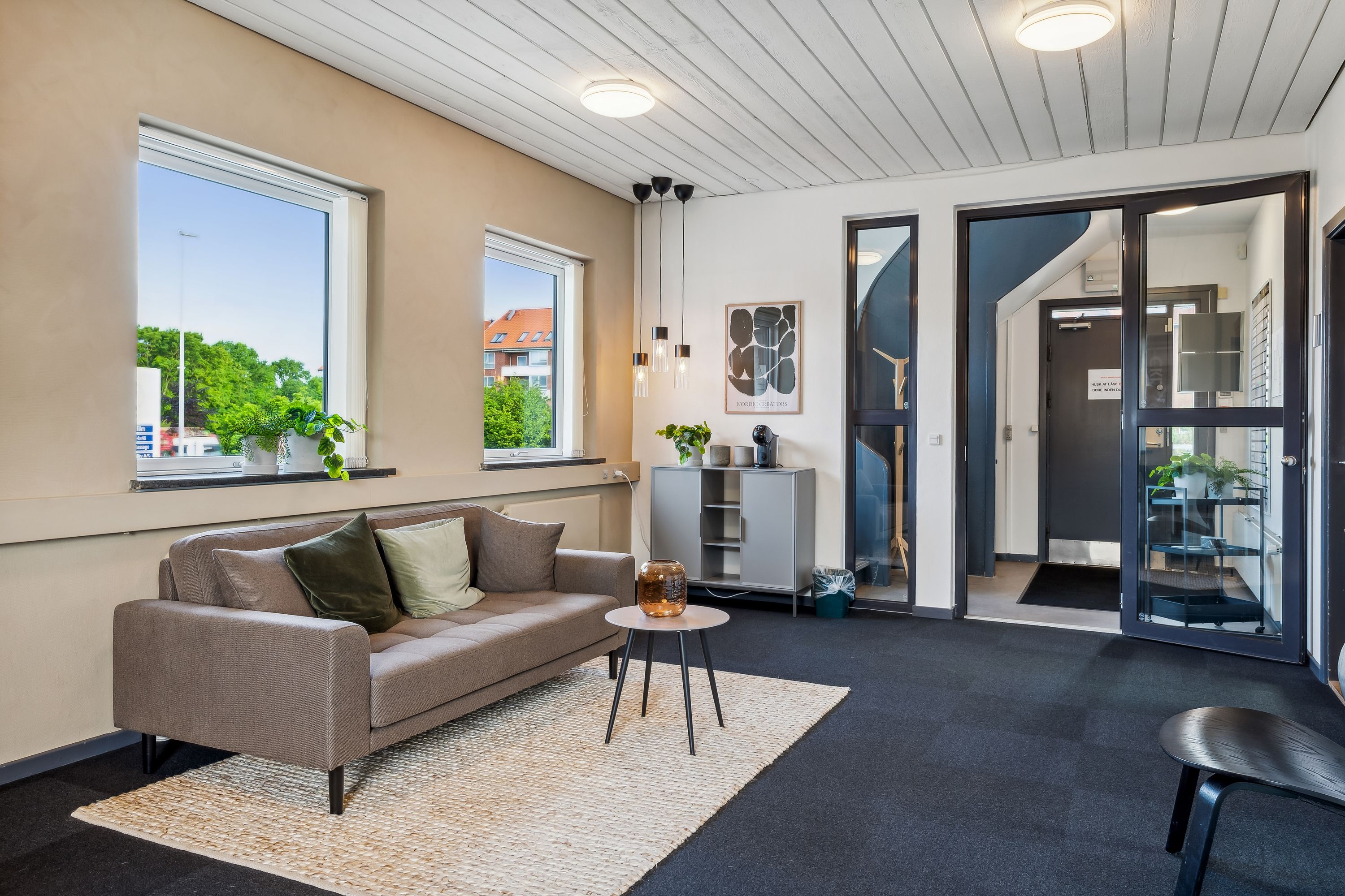 Kontorområde med sofa, stol, plante, skrivebord, vindue, glasdør og loftslys. Indeholder tekst og annoncer.